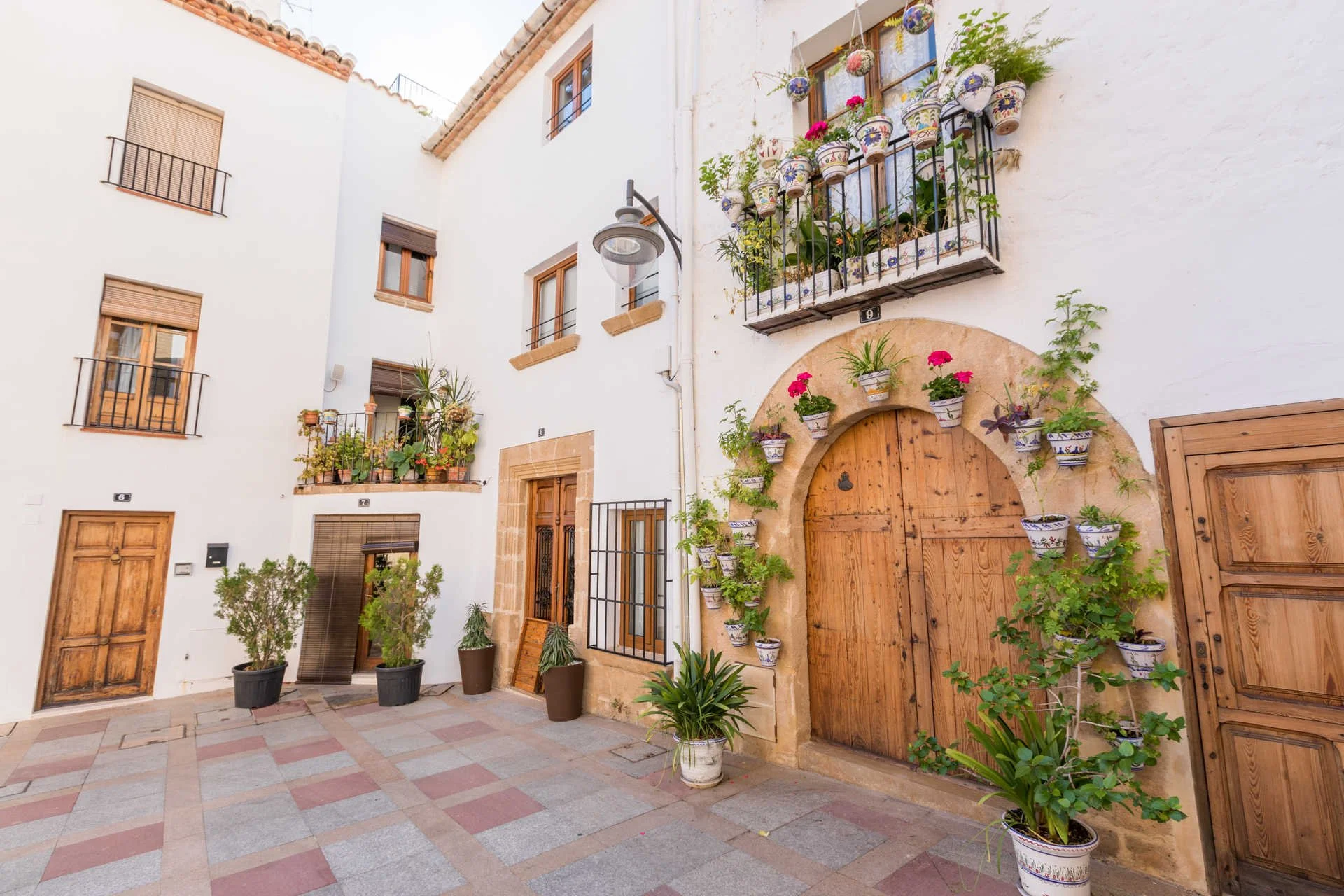 A white building with small balconies decorated with potted plants and flowers. The building has wooden doors and windows with iron railings. The courtyard has potted plants and a tiled ground.