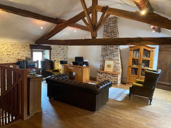 Cozy attic living room with exposed wooden beams, stone wall, brick fireplace, leather sofa, armchairs, wooden desk, bookshelf, and hardwood floors.