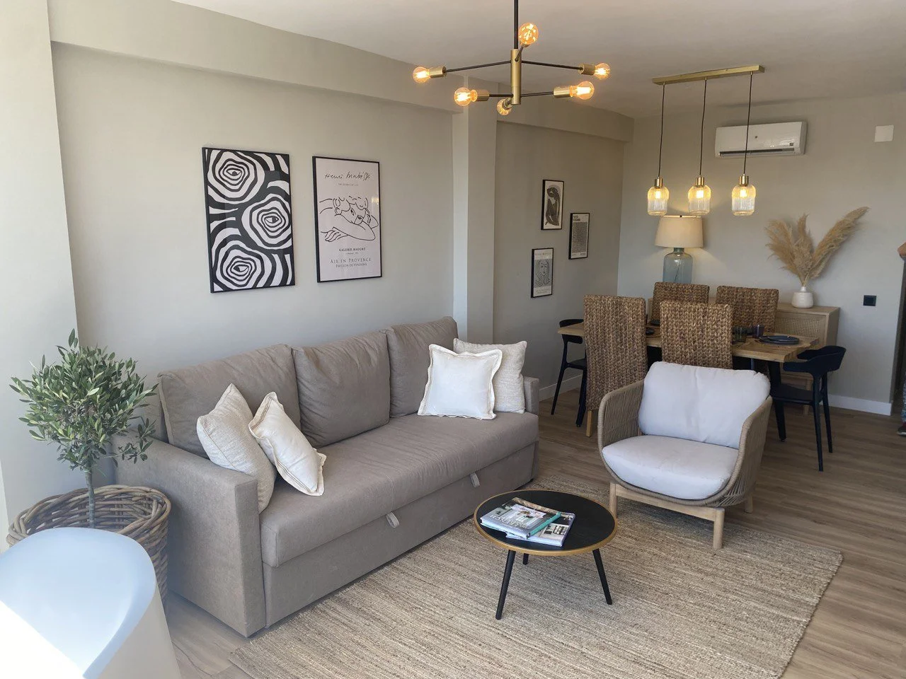 Modern living room with a beige sofa, white armchair, and a small black coffee table with magazines. There is a potted plant on the left, framed wall art, a dining area with a wooden table, wicker chairs, hanging pendant lights, and a decorative vase