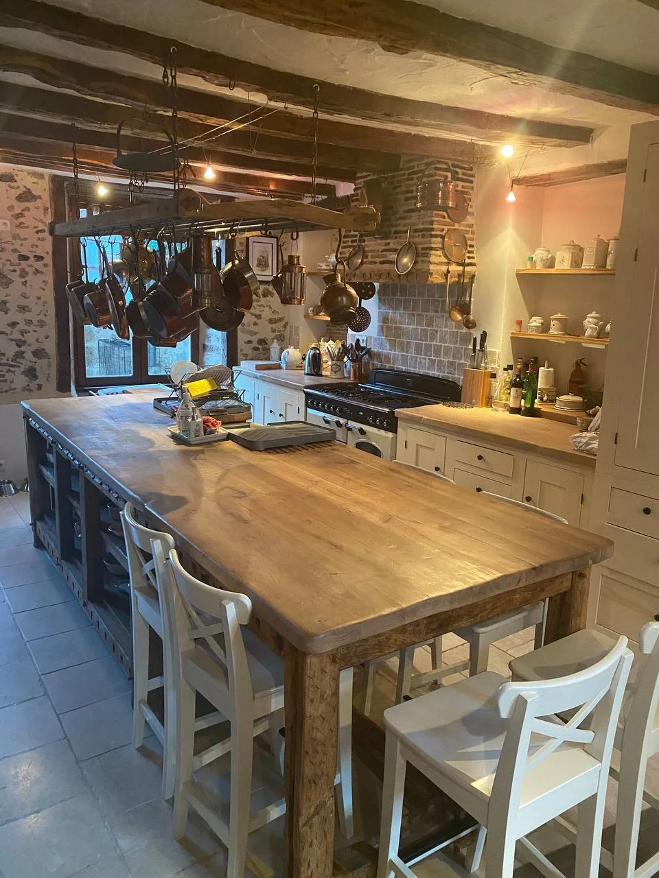 Rustic kitchen with a large wooden island, hanging pots and pans, stone and brick walls, open shelves with ceramics, and a stove with a brick chimney.
