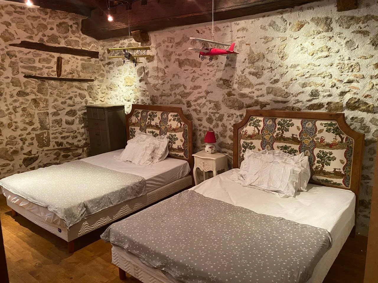 A bedroom with two beds, a stone wall, and hanging model airplanes.