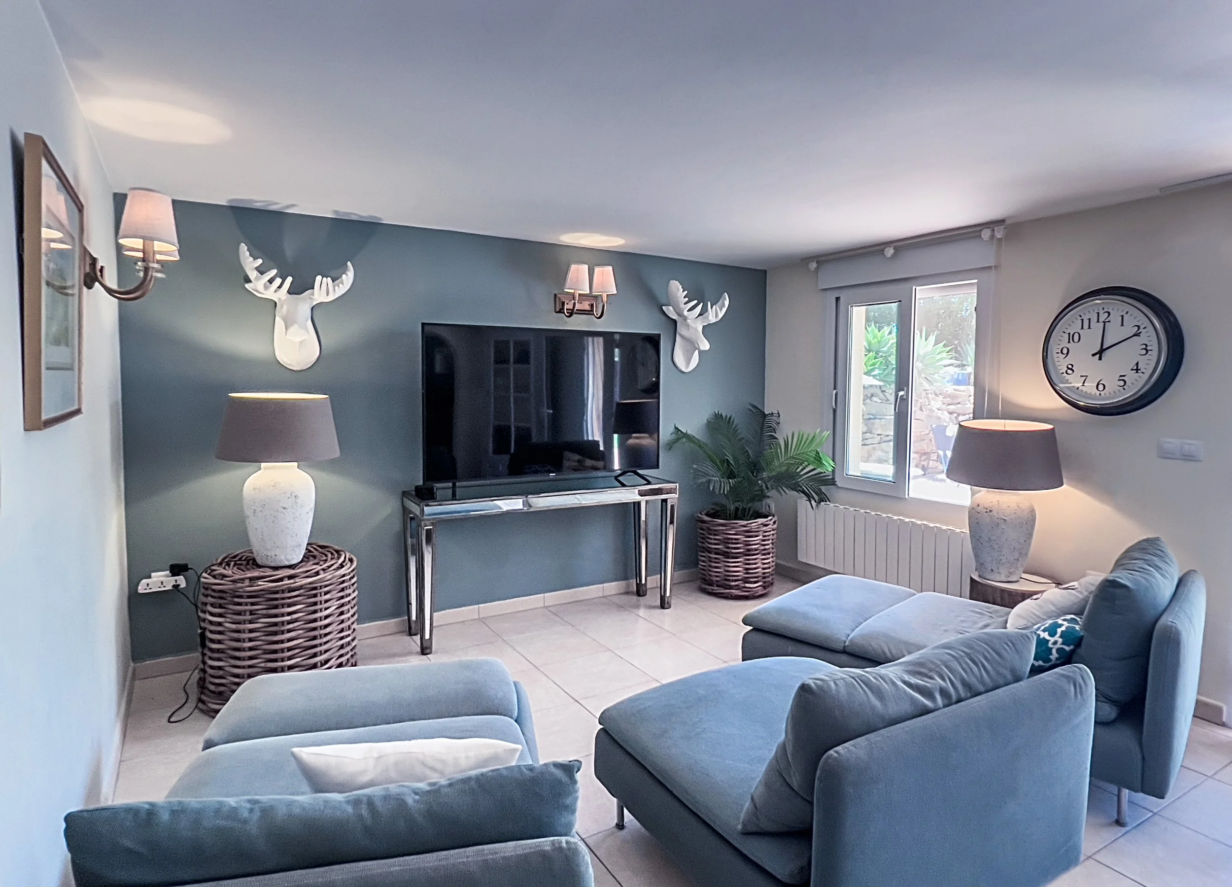 Living room with blue walls, wall-mounted decor including white deer heads, a large flat-screen TV, two table lamps with beige shades, a potted plant, a window with outdoor view, a large clock, and blue furniture including a sofa and ottoman.