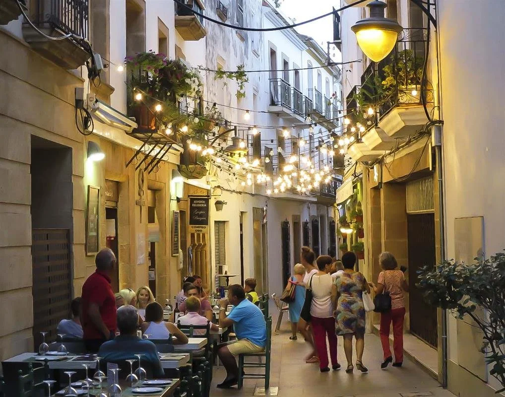 A narrow outdoor street with outdoor seating at a restaurant, string lights overhead, people dining and walking, and buildings with balconies and flowers.
