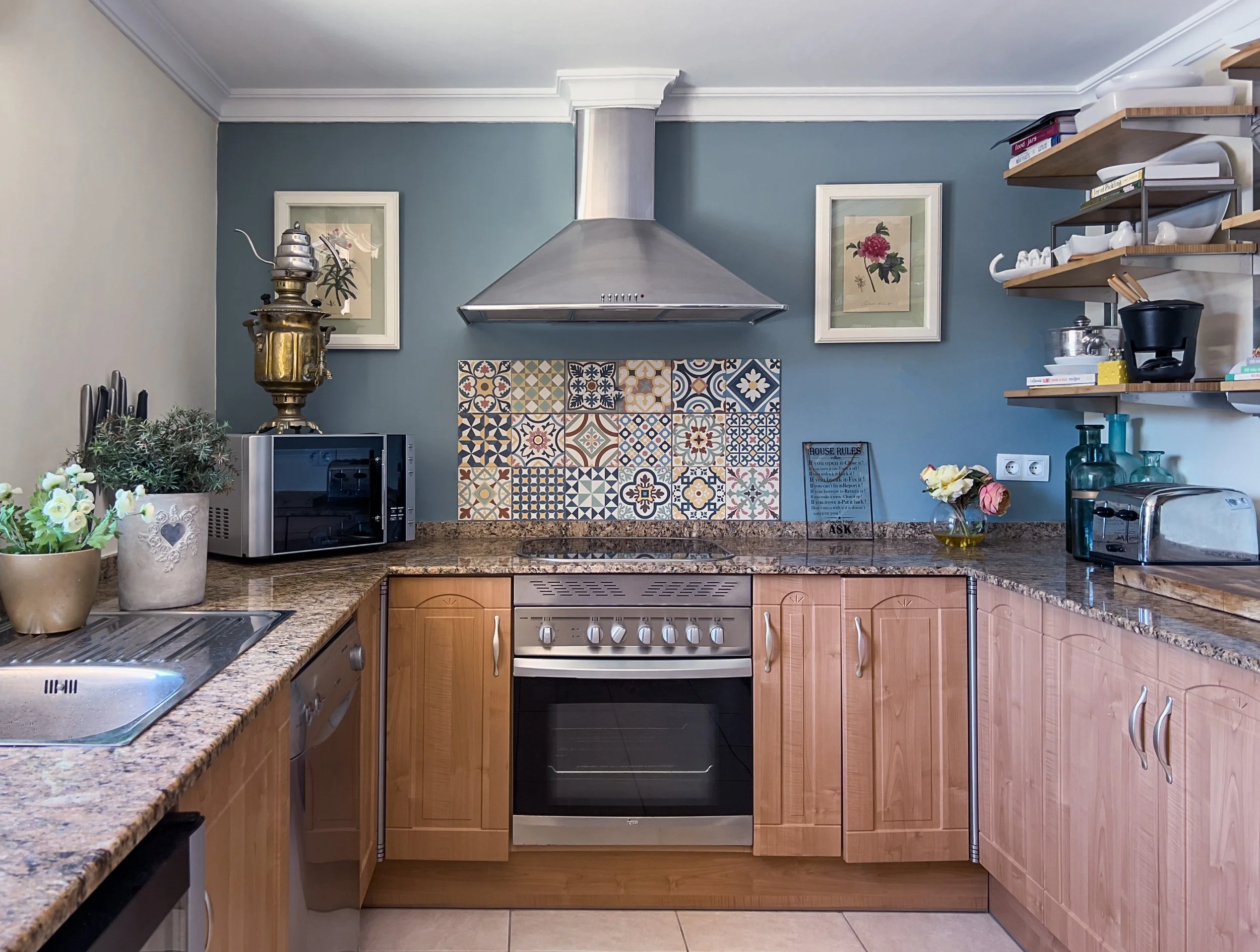 Kitchen with granite countertops, light wood cabinets, a stainless steel oven and stove, a stainless steel range hood, a microwave, open shelves with dishes, a small flower arrangement, plants, and decorative tiles on a blue wall.