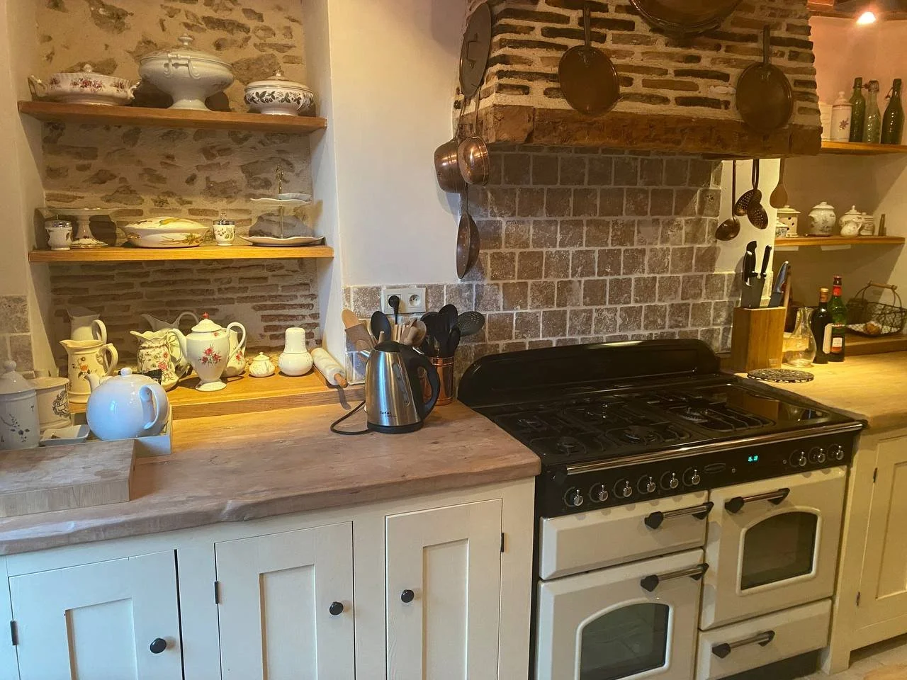 Kitchen with brick wall and open shelves displaying teapots, dishes, and glassware. A vintage stove with pots hanging above, alongside a countertop with a kettle, utensils, and bottles.