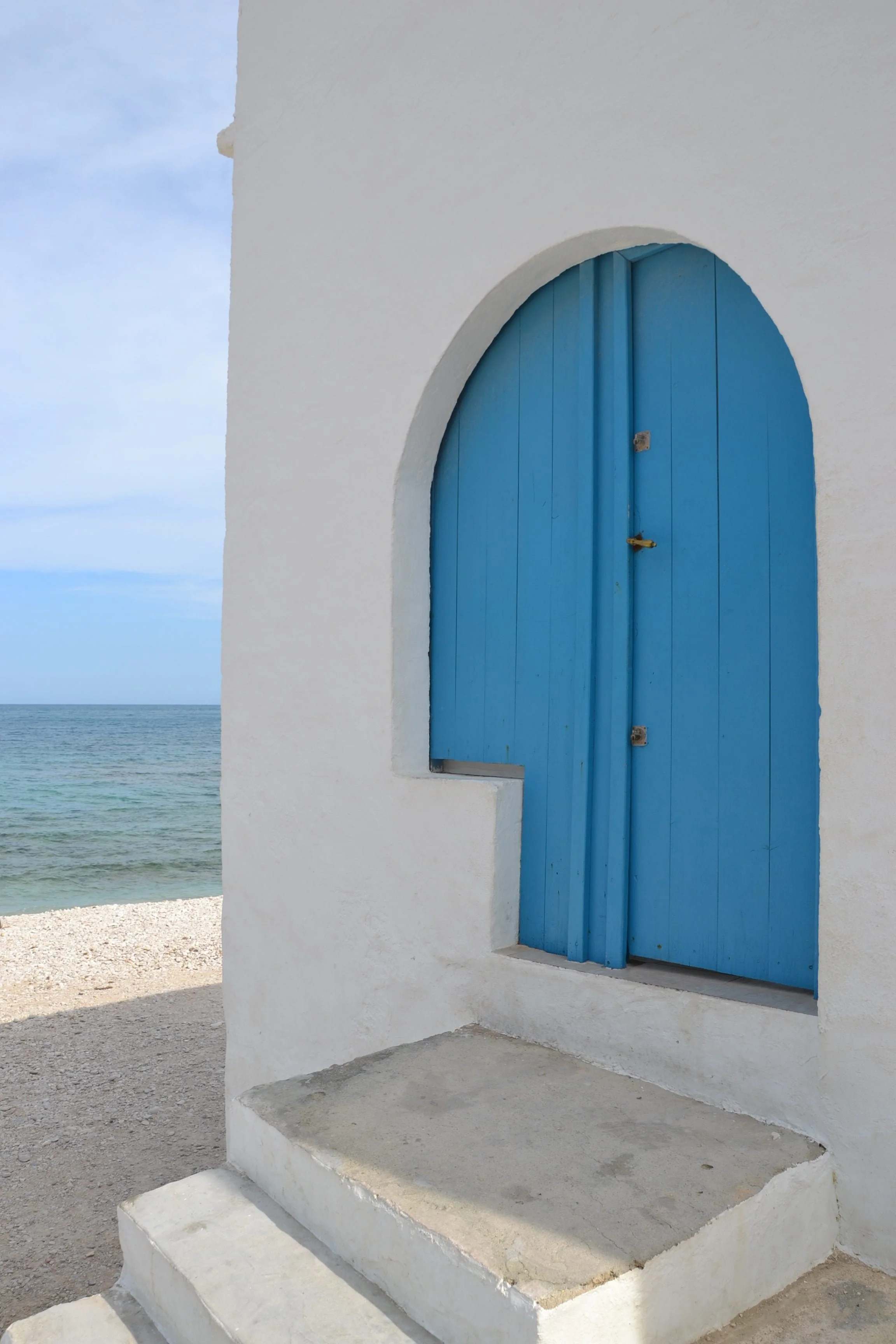 White building with a curved blue door, steps leading up to it, with the ocean in the background under a partly cloudy sky.