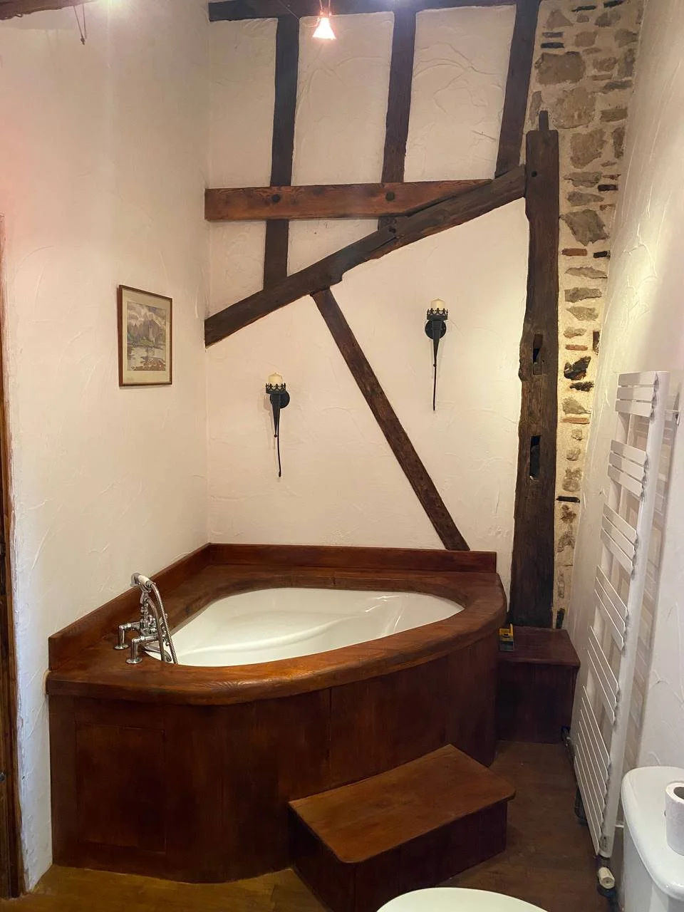 A corner bathroom with a built-in wooden tub, rustic wooden beams on the wall, a small wooden stool, and wall-mounted lights. There is also a framed picture and a towel warmer.