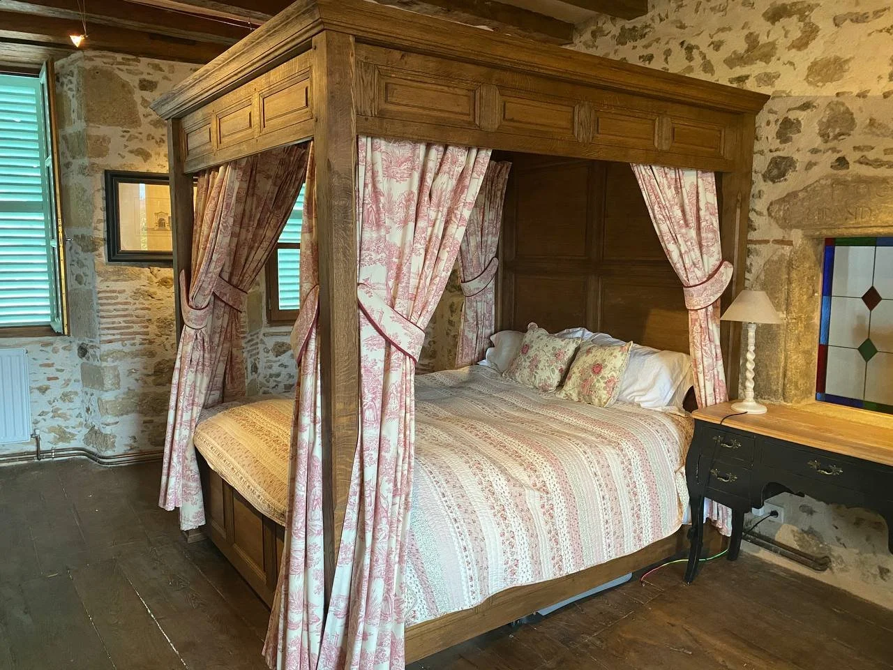 A rustic bedroom with a four-poster wooden bed with pink and white patterned curtains and bedding, set against stone walls, with green shuttered windows and a small black desk with a stained glass window beside it.