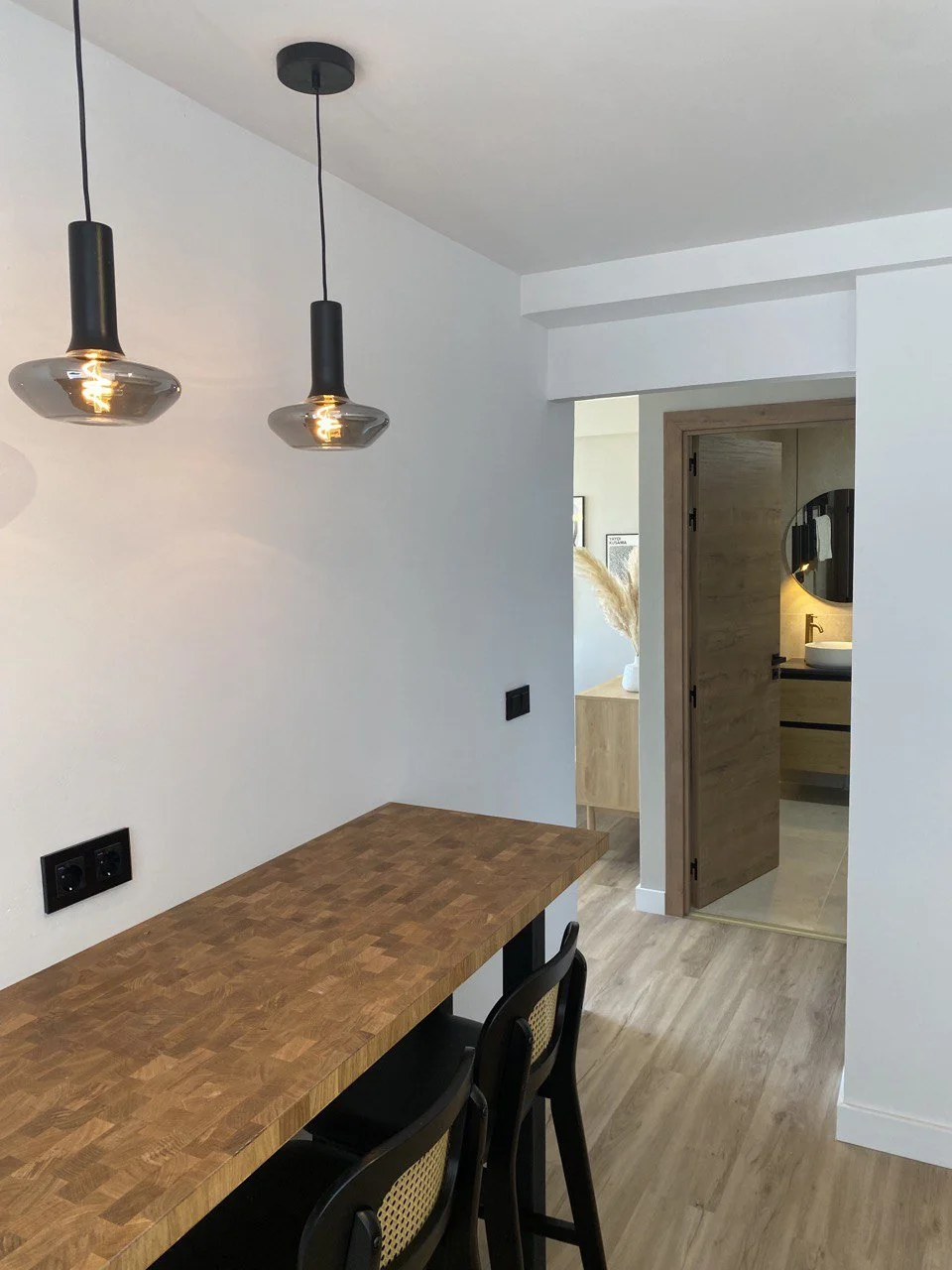 Interior view of a dining area with a wooden table and two black chairs, pendant lights hanging from the ceiling, and a doorway leading to a bathroom with a mirror and sink visible.
