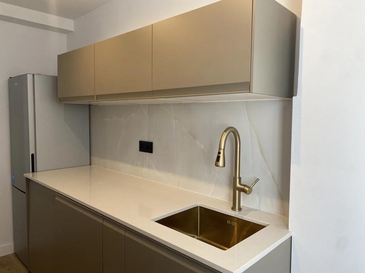 Modern kitchen with beige upper cabinets, a white countertop, a gold sink, and a gold faucet. There is a black power outlet on the backsplash and a silver refrigerator on the left side.