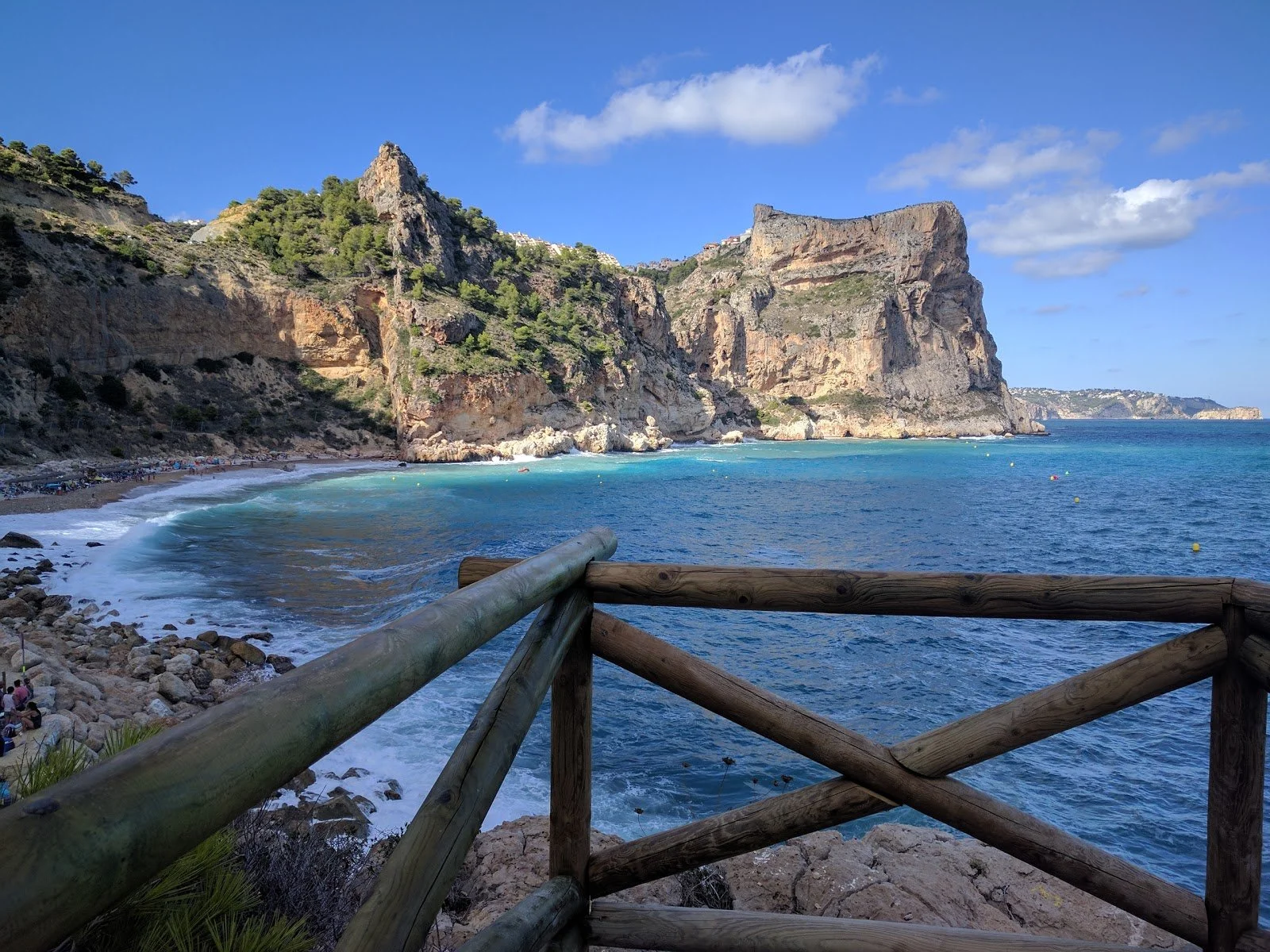 Scenic coastal view with rocky cliffs, blue ocean, and a viewing platform with a wooden railing overlooking the beach and cliffs.