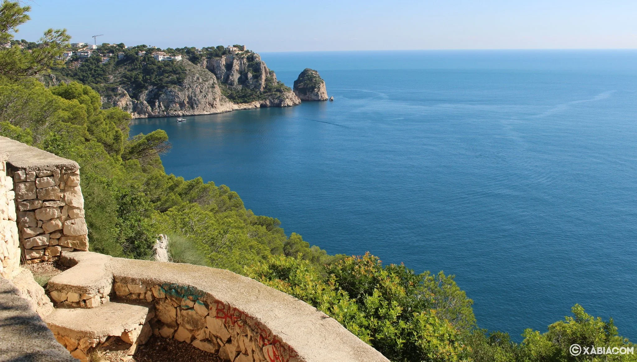 Scenic view of a coastal landscape with clear blue water, green trees, rocky cliffs, and a few white buildings on a hillside in the distance.