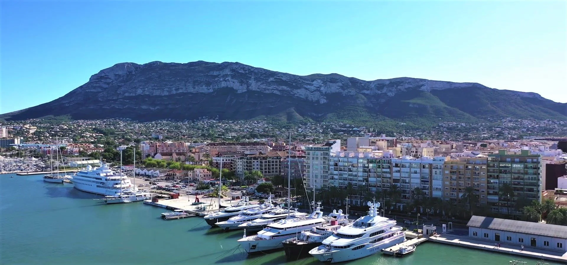 View of a marina with several yachts docked and a cityscape, with a mountain in the background under a clear blue sky.