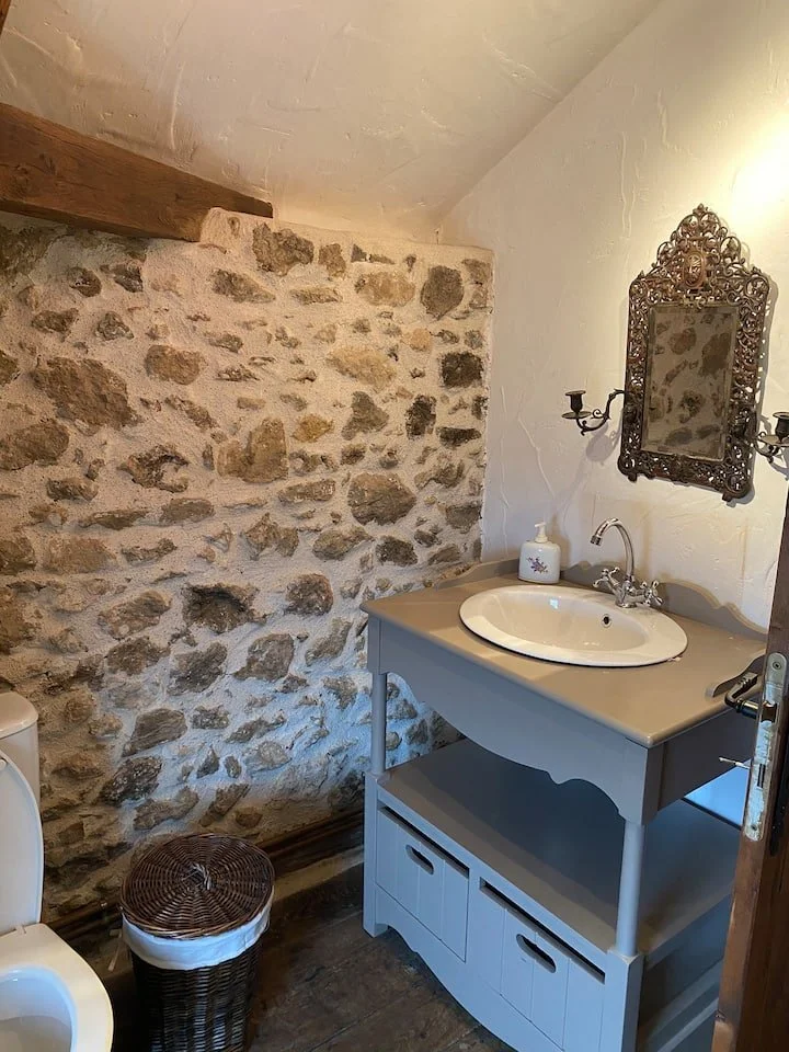 A rustic bathroom corner with a stone wall, wooden beam ceiling, vintage mirror, basin sink on a white vanity, soap dispenser, and a wicker laundry basket.