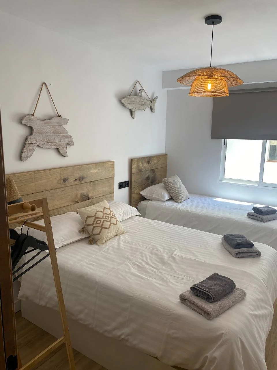 Bedroom with two beds, wooden headboards, gray and beige towels on beds, decorative pillows, wall-mounted wooden fish decorations, window with gray roller shade, and wicker pendant light.
