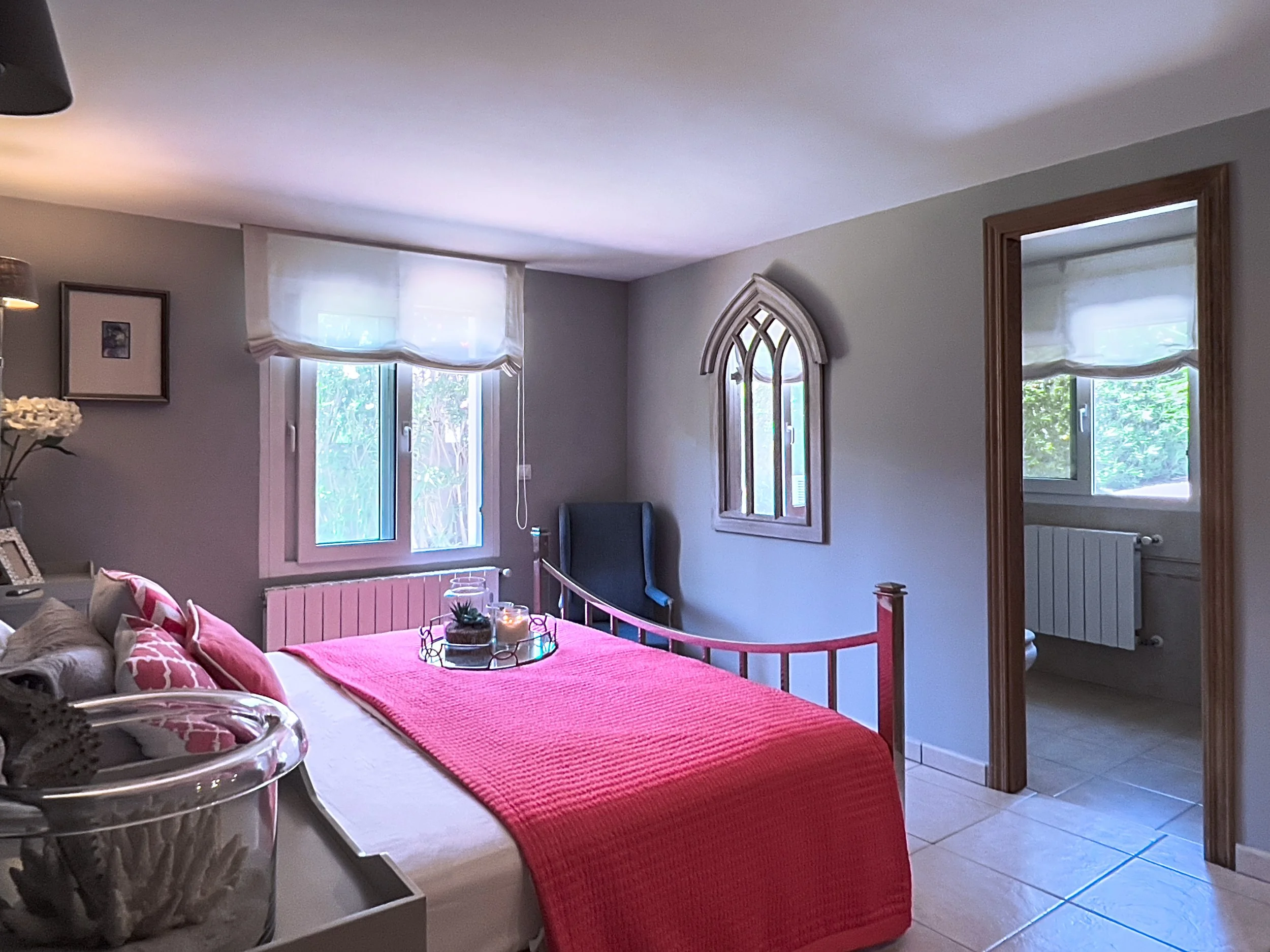 A bedroom with a bed covered in a pink blanket and pillows, a window with white sheer curtains, and an adjoining bathroom with a white toilet visible through an open doorway.
