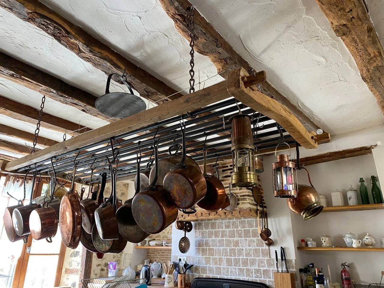 Hanging copper and metal pots and pans in a rustic kitchen with exposed wooden beams and brick wall.