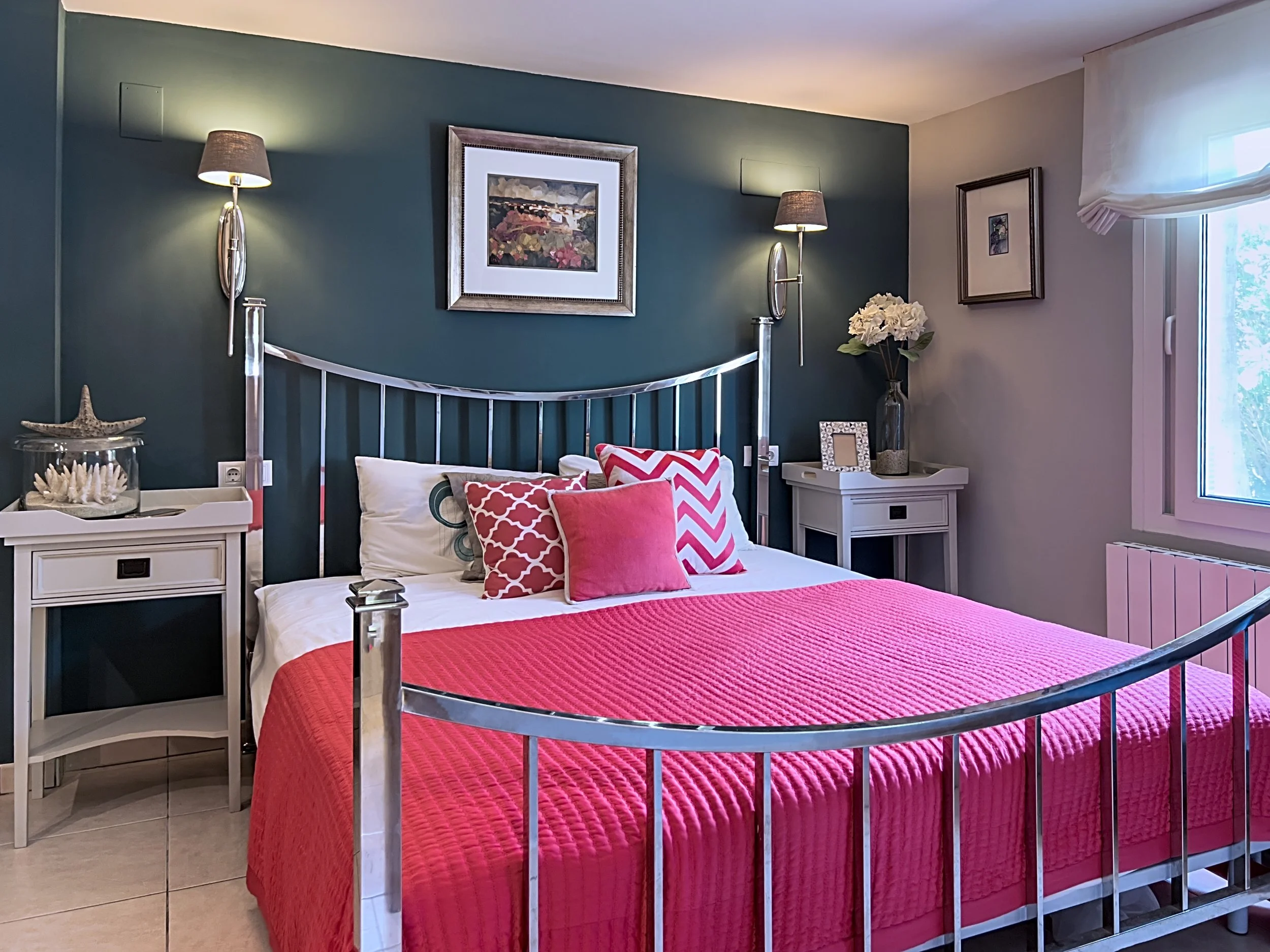 Bedroom with a metal bed frame, pink bedspread, and throw pillows, including chevron and solid designs. Two white nightstands with lamps and decor, a window with white curtains, wall art, and a teal accent wall.