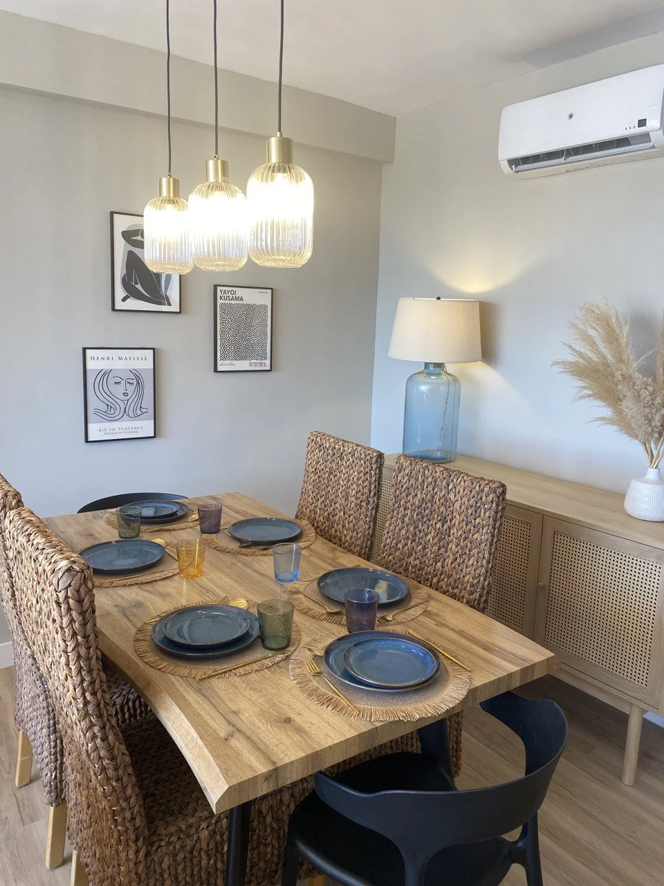 A dining room featuring a wooden table set with blue plates, woven placemats, and multicolored glasses, surrounded by rattan chairs. The decor includes framed art on the wall, a large glass lamp on a sideboard, and a white vase with pampas grass. An
