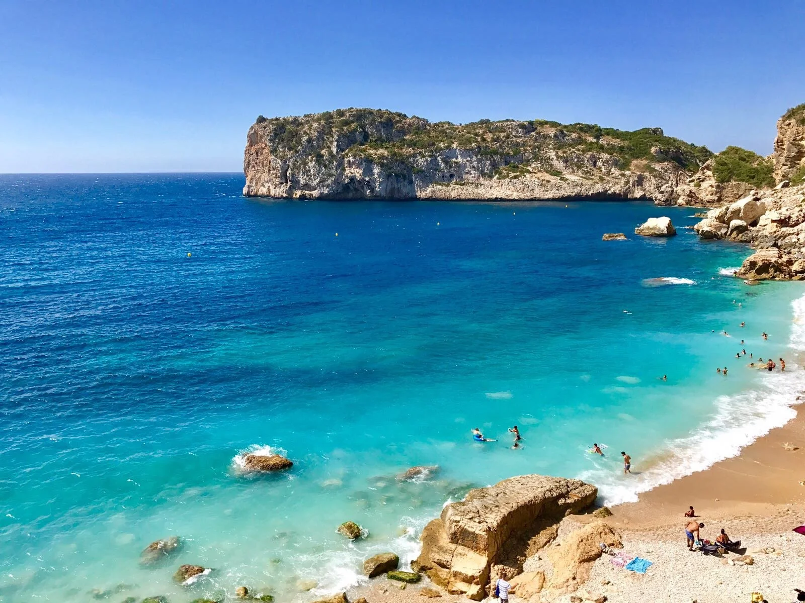 A scenic beach with turquoise water, large rocks, and cliffs in the background, with people swimming and relaxing on the shore under a clear blue sky.