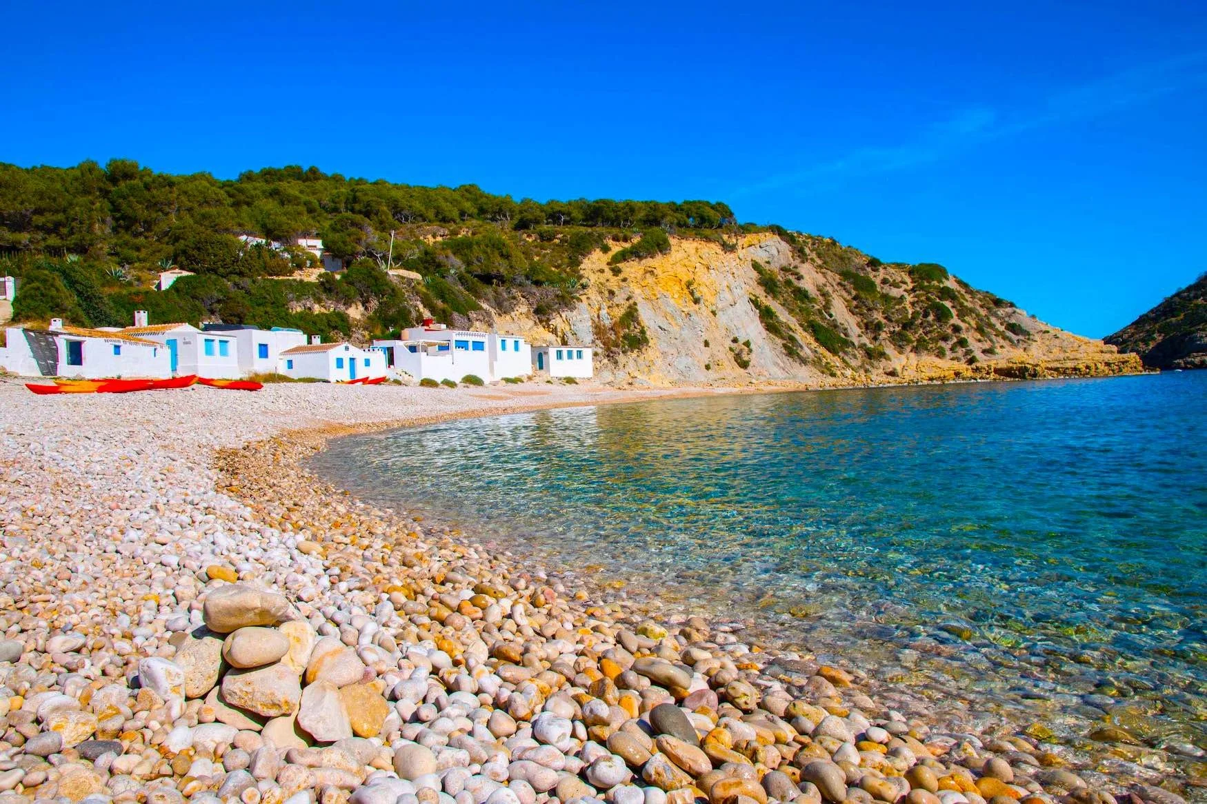 Pebble beach with white houses and blue windows along the shoreline, hills covered in greenery, clear blue sky, and calm water.