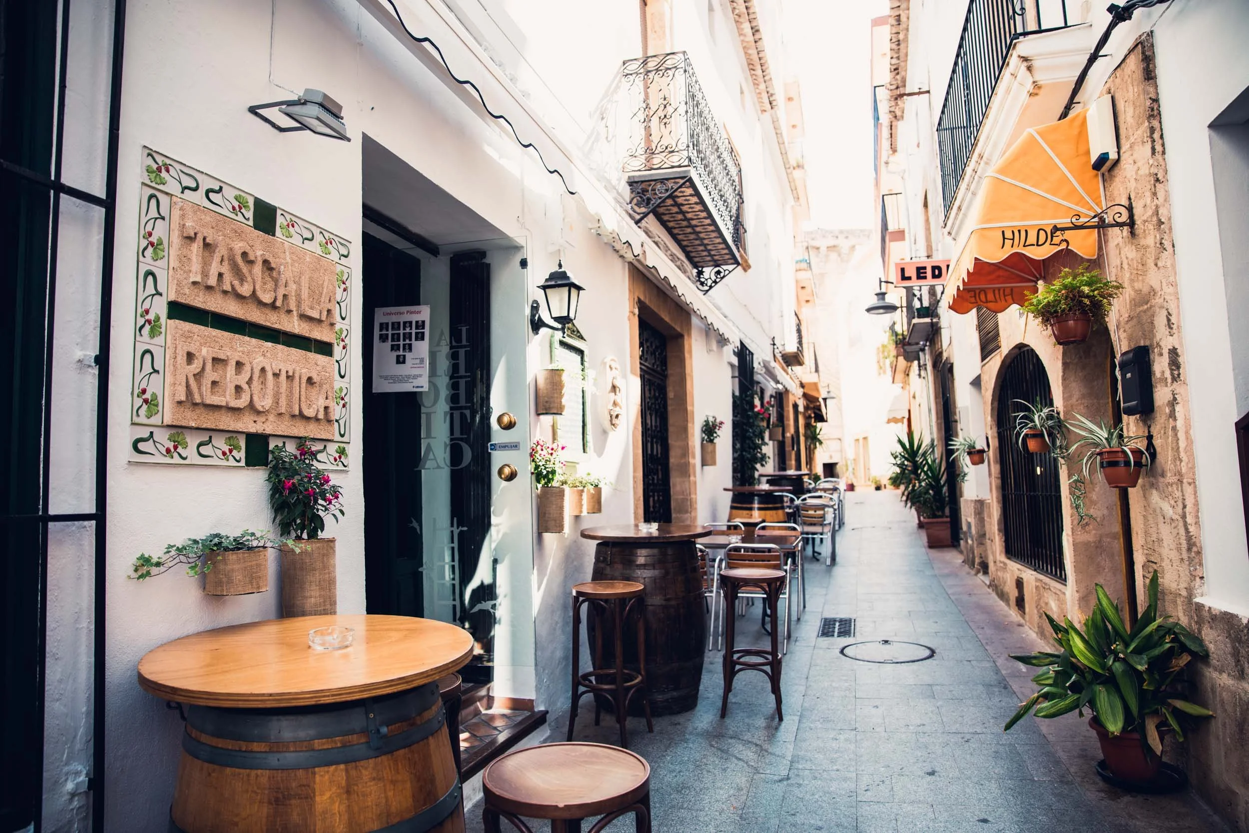 Empty outdoor alleyway in a European city with small cafes, potted plants, and hanging flower baskets, sunlit and quiet.