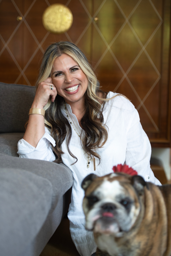 Woman with long wavy hair, smiling and sitting on the floor next to a bulldog, with a modern wooden wall and spherical hanging light in the background.