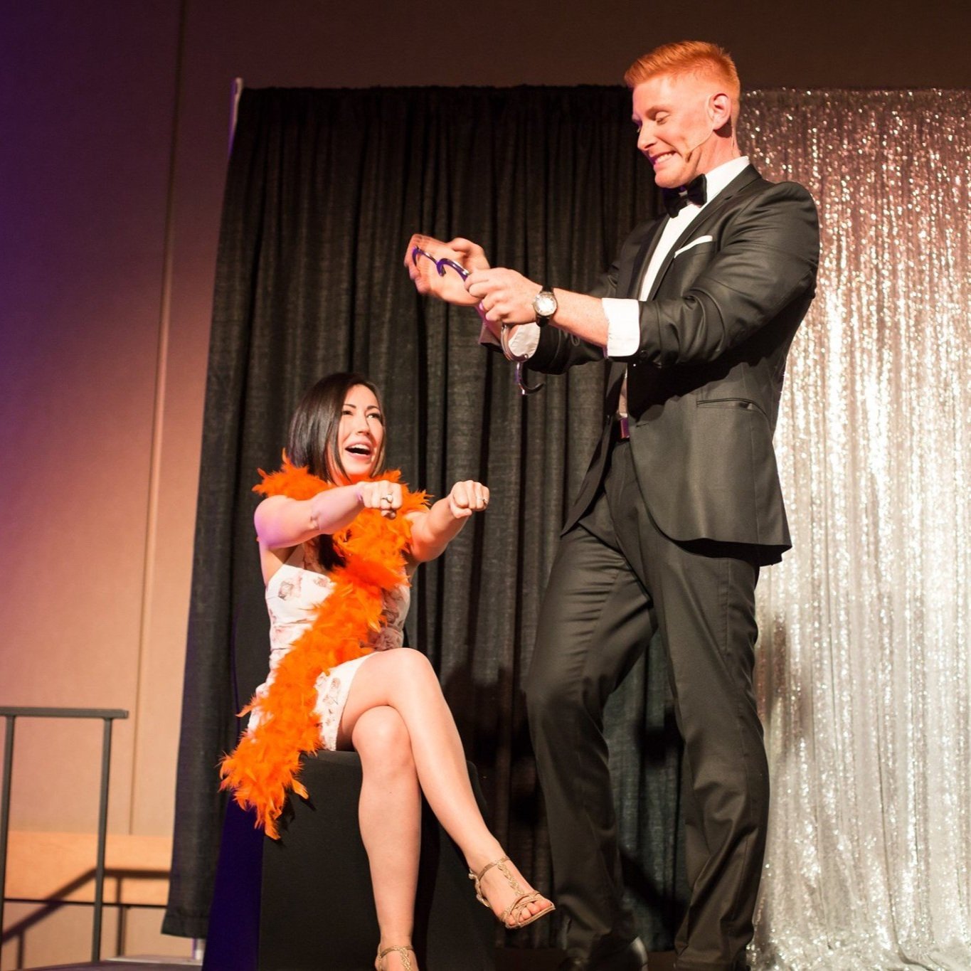  Matt Gore, a magician, is seen in a tuxedo holding a handcuff, accompanied by a woman in a black dress wearing an orange feather, holding her hands high for a magic show 