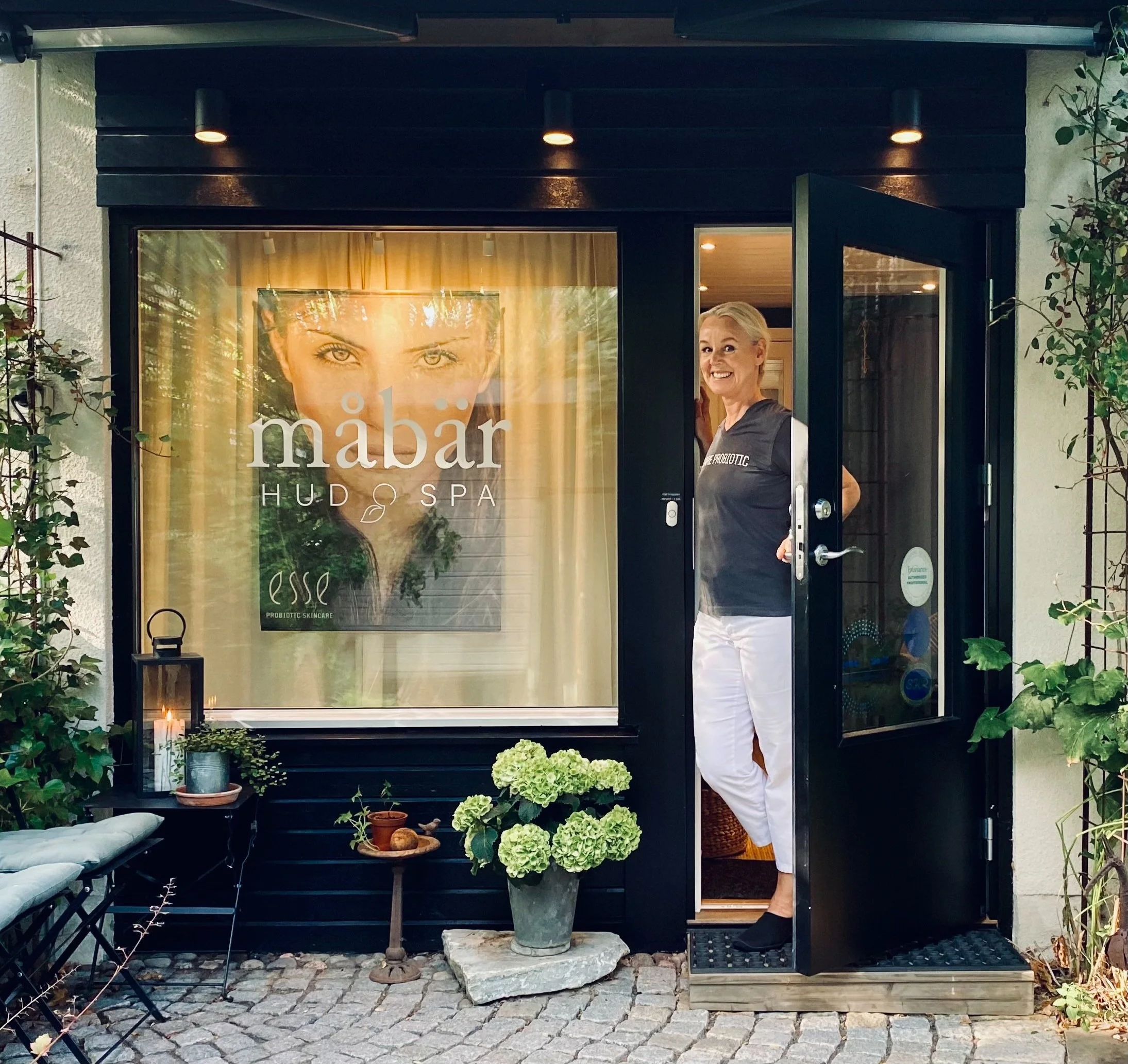 En inbjudande exteriörbild av Måbärs salong med Anette vid dörren.