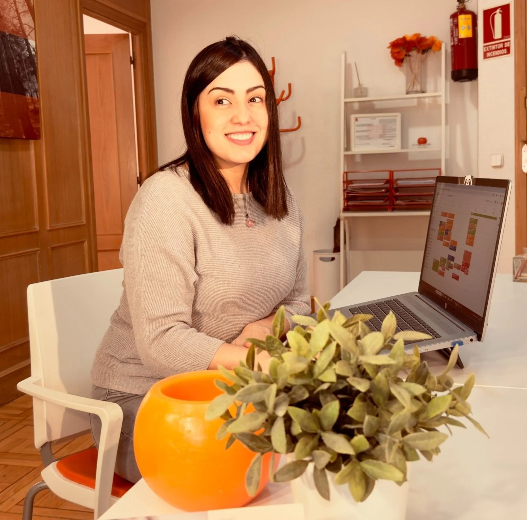 Mujer sonriendo en una oficina, sentada frente a una computadora portátil, con plantas y objetos decorativos en el fondo.