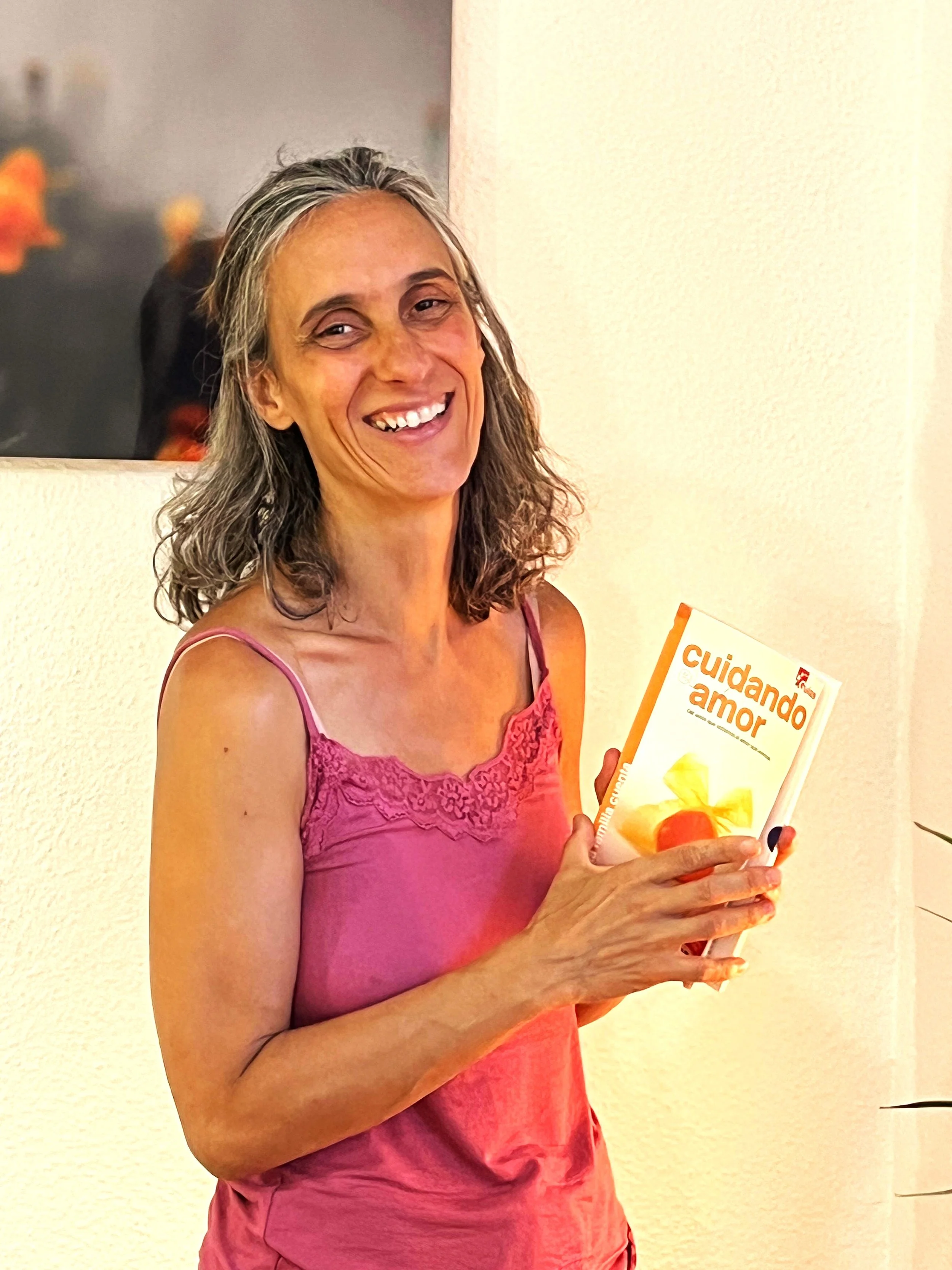 Mujer de cabello canoso con sonrisa sosteniendo un libro que dice 'Cuidando amor' en un fondo interior de casa.