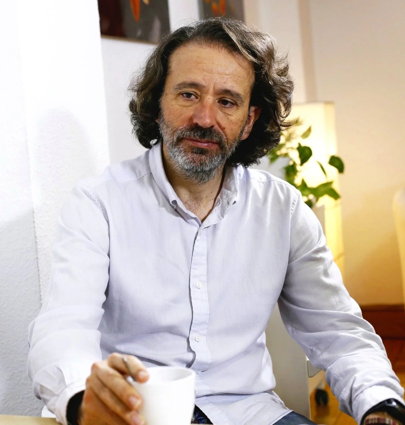 Hombre de cabello largo y barba gris, sentado en una habitación, sosteniendo una taza blanca.