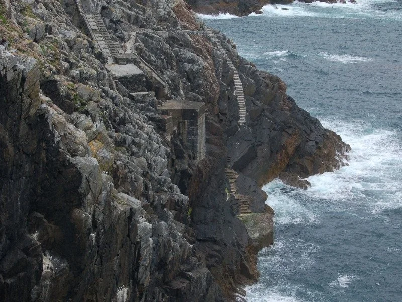 Steep cliffs above rough sea. Two sets of steps lead down the side of the rocks. One goes to a platform about half way down, the other leads to a landing platform just above sea level.