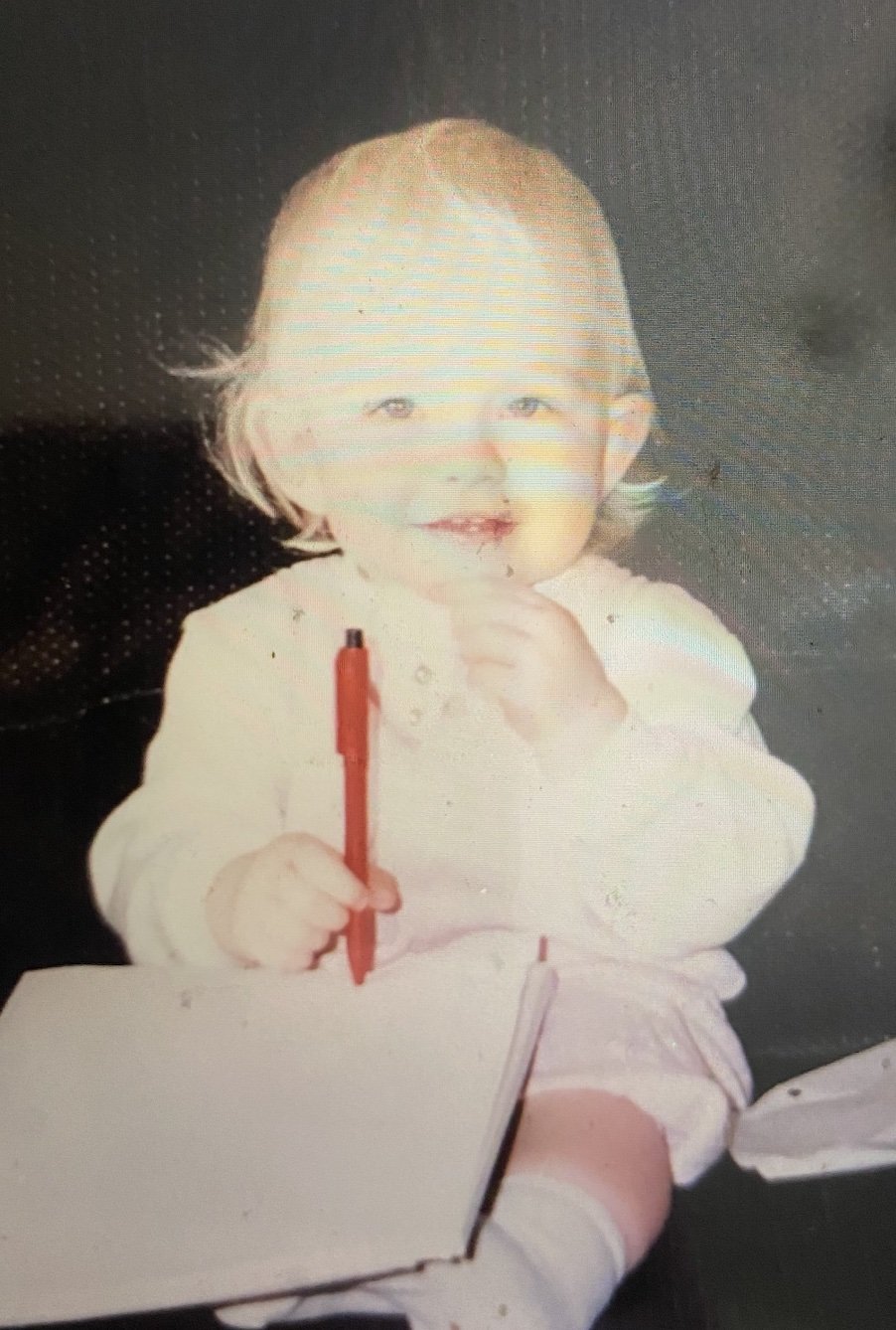 Me at the age of around 18 months, holding a pen with paper in her lap.