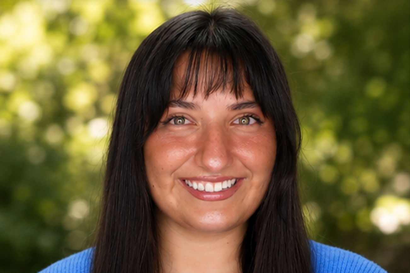 Smiling woman with long dark hair wearing a bright blue cardigan, standing against a plain white background.