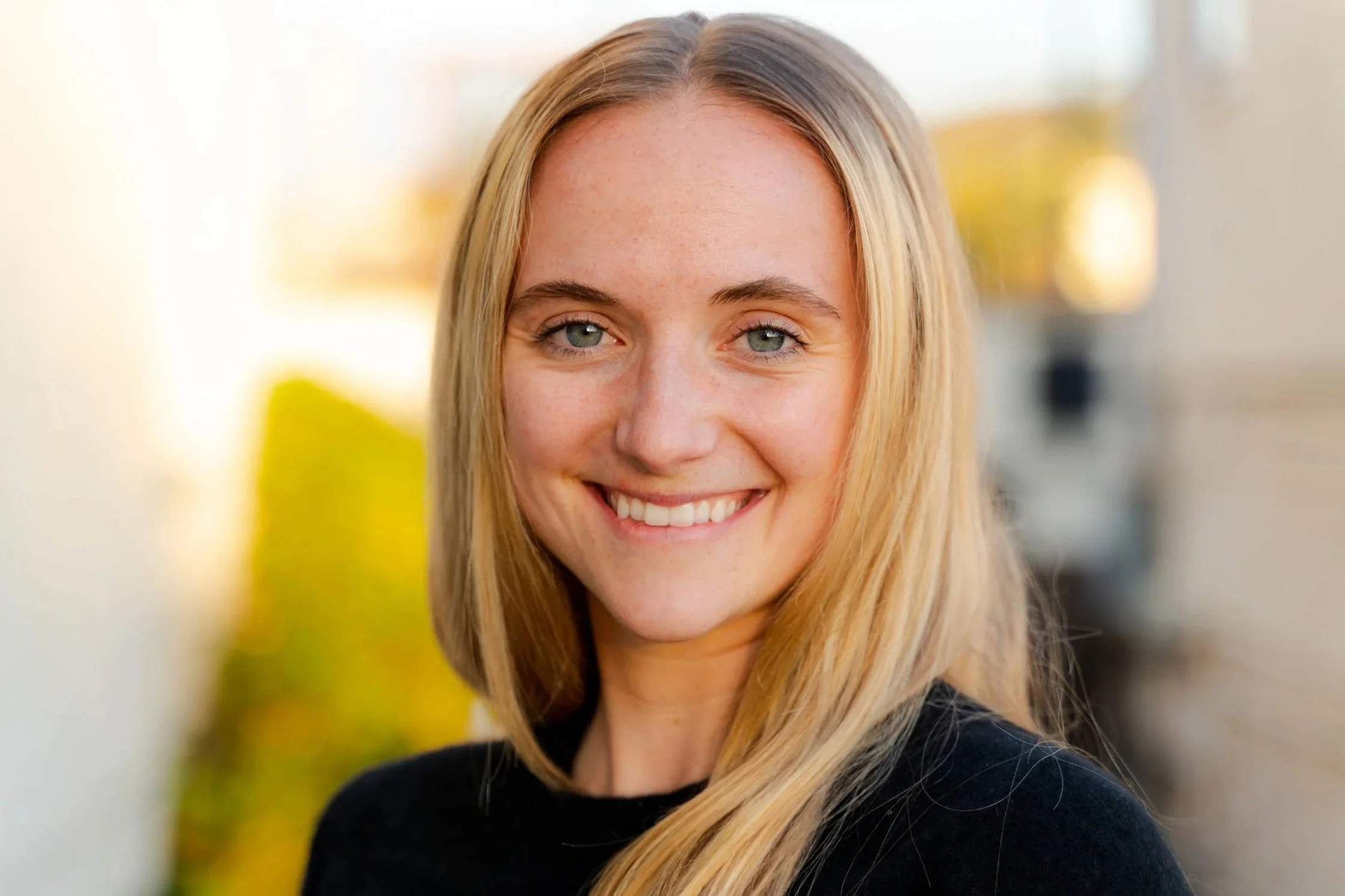 Woman smiling outdoors, wearing a black top with long blonde hair, blurred background.