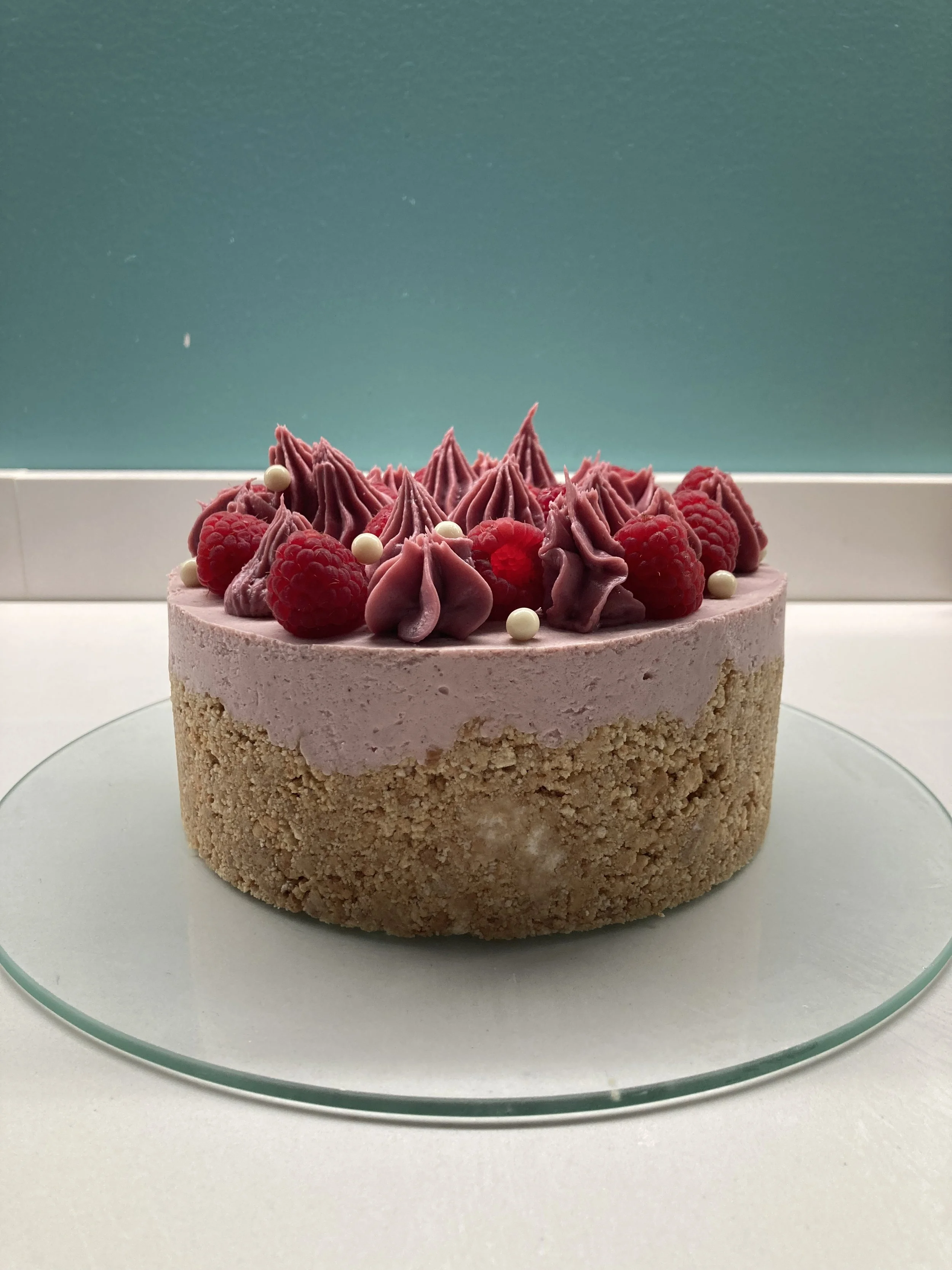 Cheesecake con base di biscotto, copertura di crema di lamponi, decorata con lamponi freschi, cioccolato bianco e ganache al cioccolato rosa Callebaut