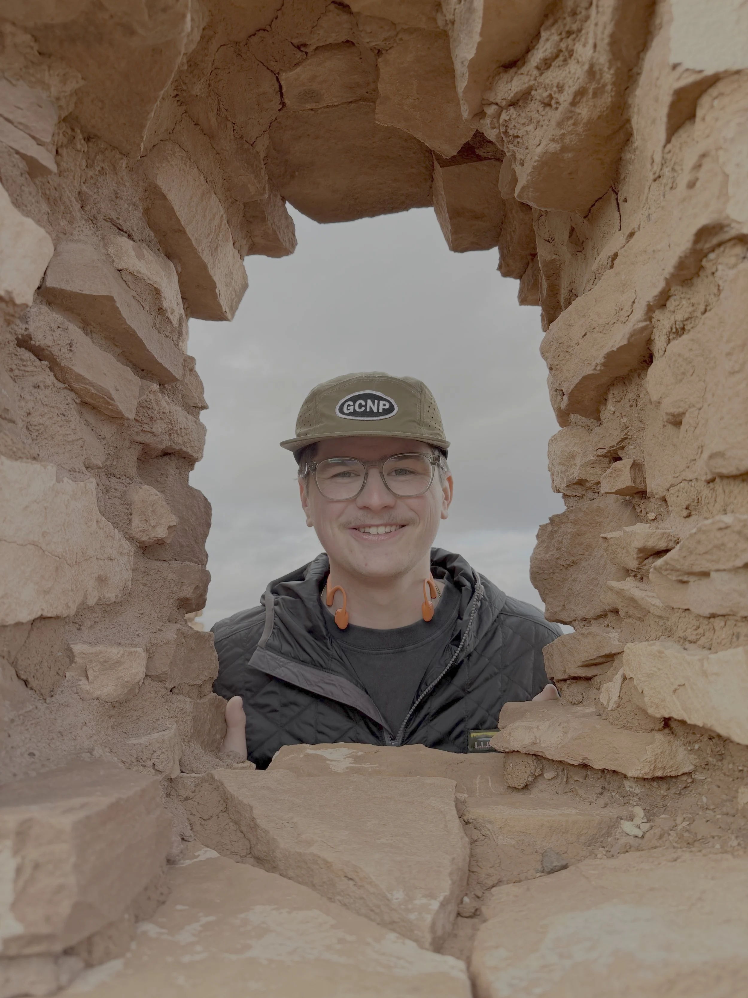 A person smiling through a window made of old red bricks, wearing glasses, a tan cap with a logo, and a black jacket.