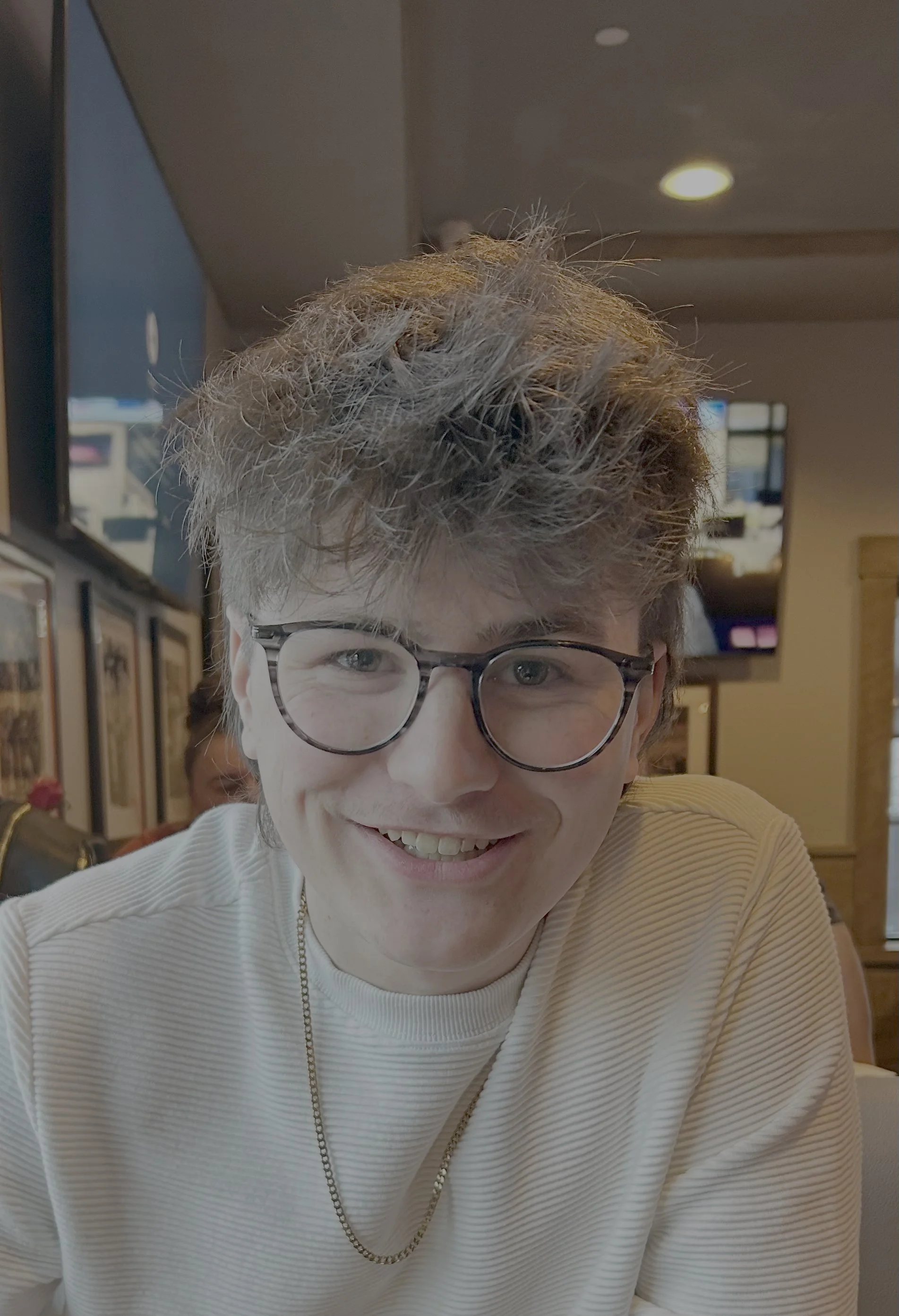 Close-up of a young person with curly, tousled hair wearing glasses and a white, ribbed shirt, smiling in a restaurant or cafe.