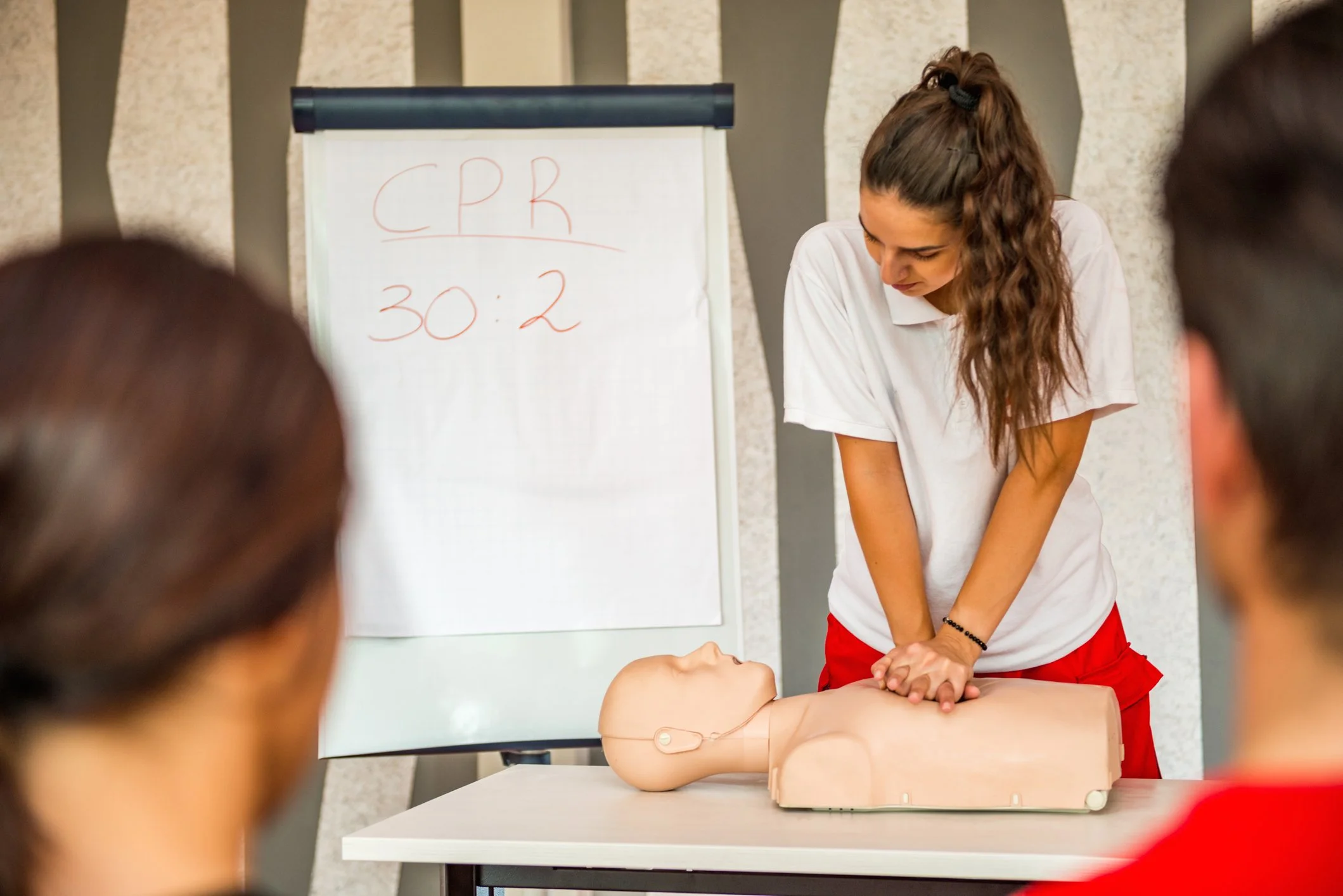 A woman demonstrating CPR on a mannequin during a training session, with a whiteboard in the background displaying CPR ratio 30:2.