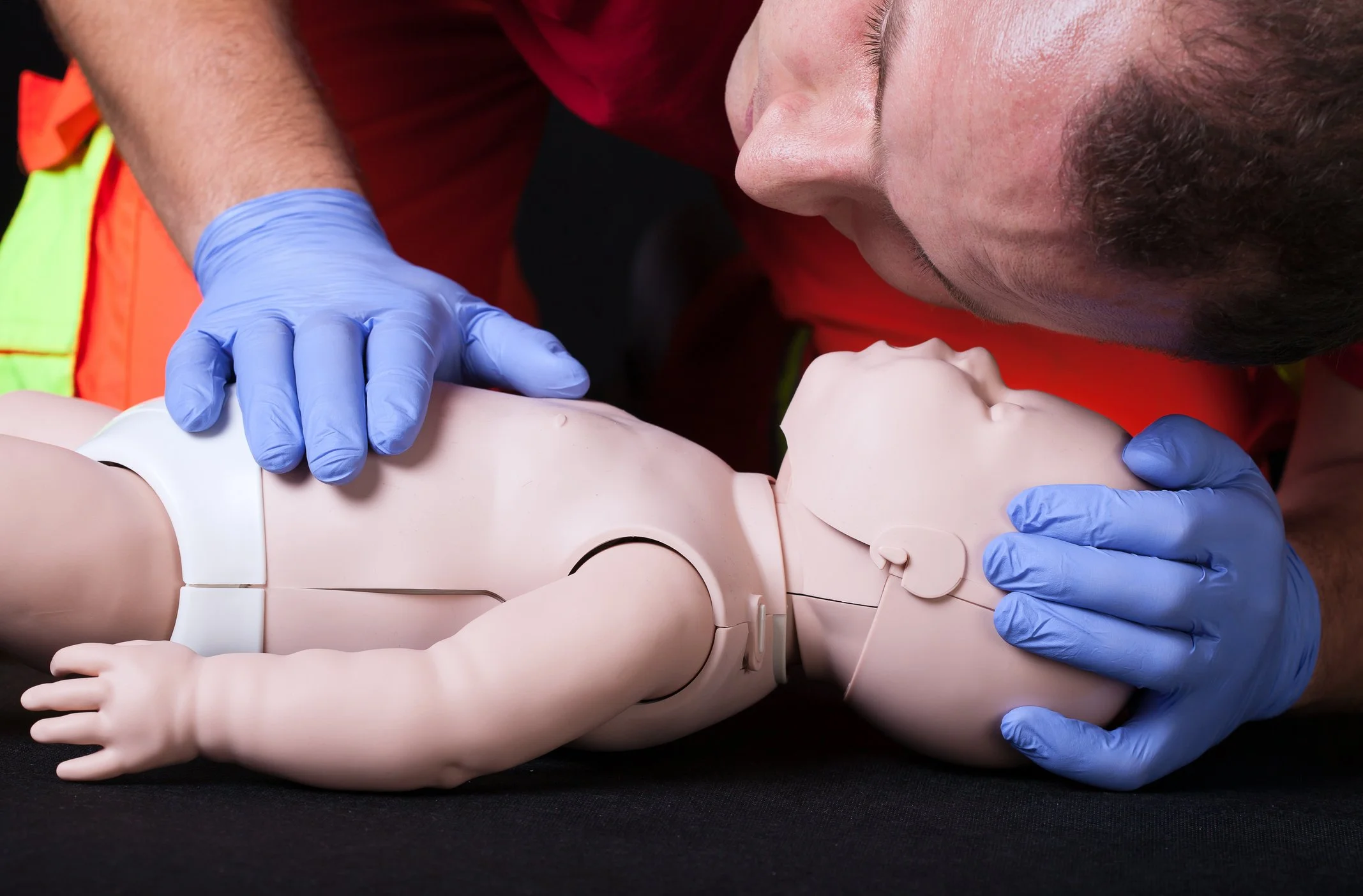 Person performing CPR on an infant mannequin, wearing blue gloves, lying on black surface.