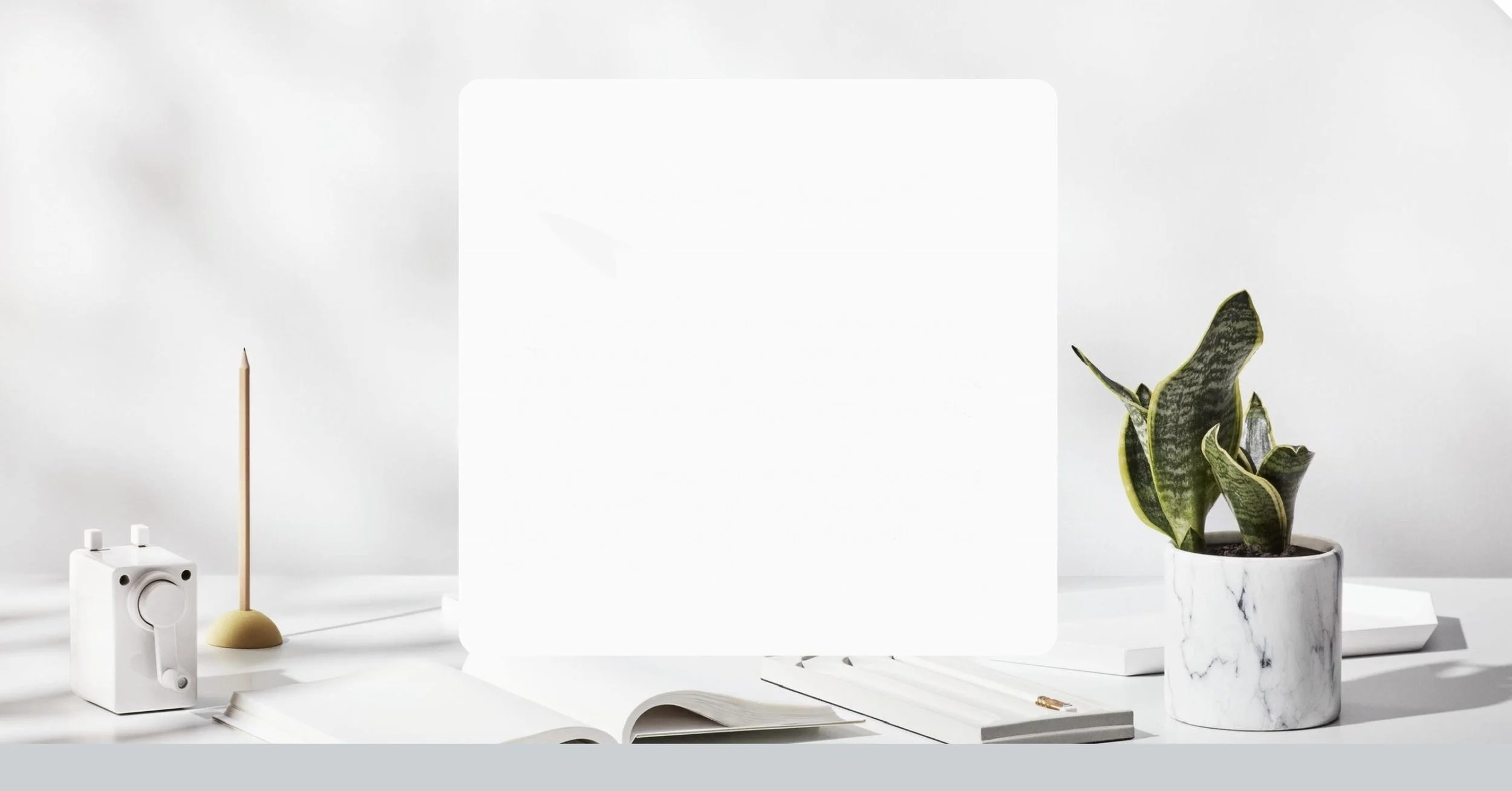Minimalist workspace with a white potted snake plant, an open white notebook, a white camera-shaped device, pencils, and a white tray, set against a light background.