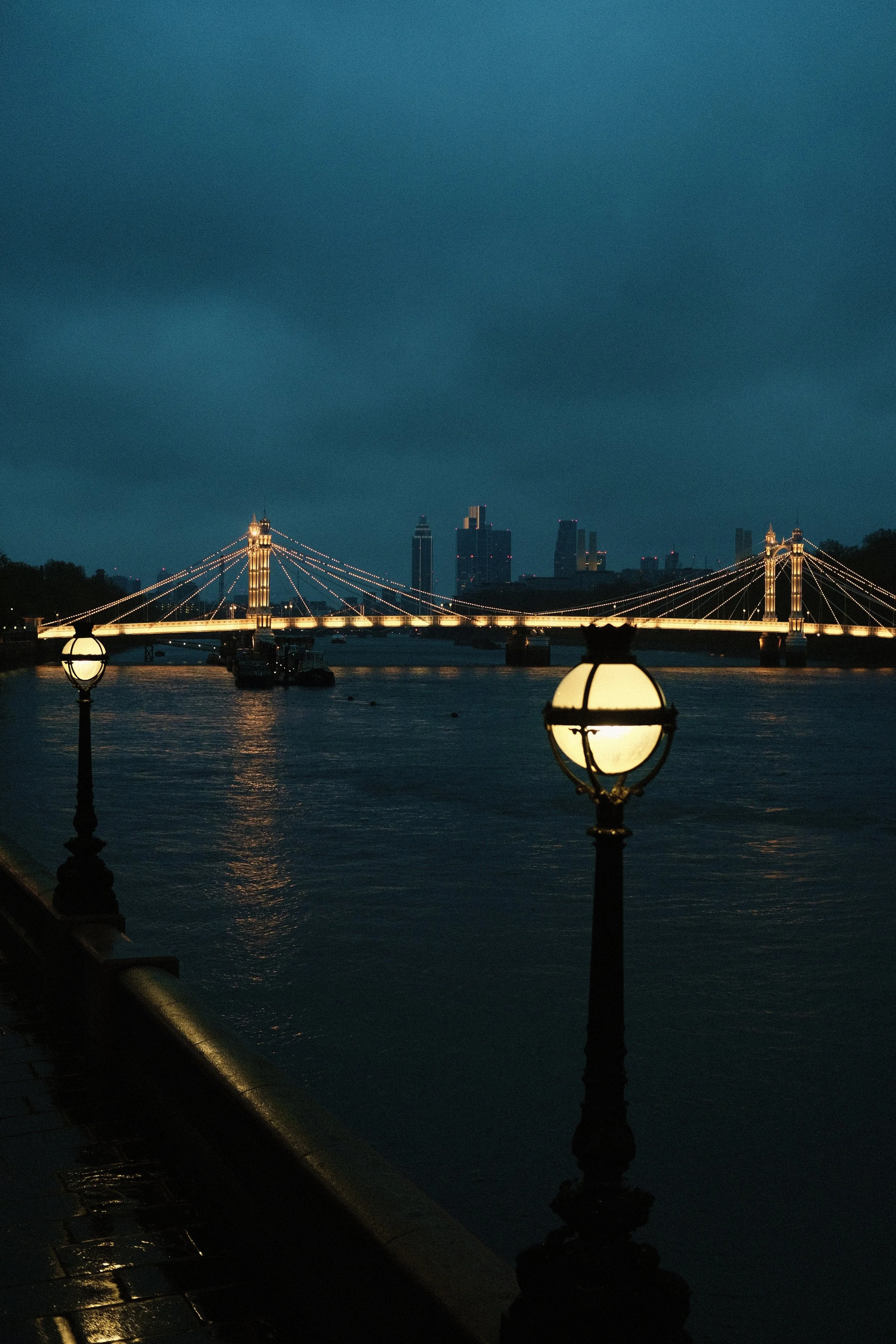 Illuminated bridge over a river at night with street lamps in the foreground and city skyline in the background.
