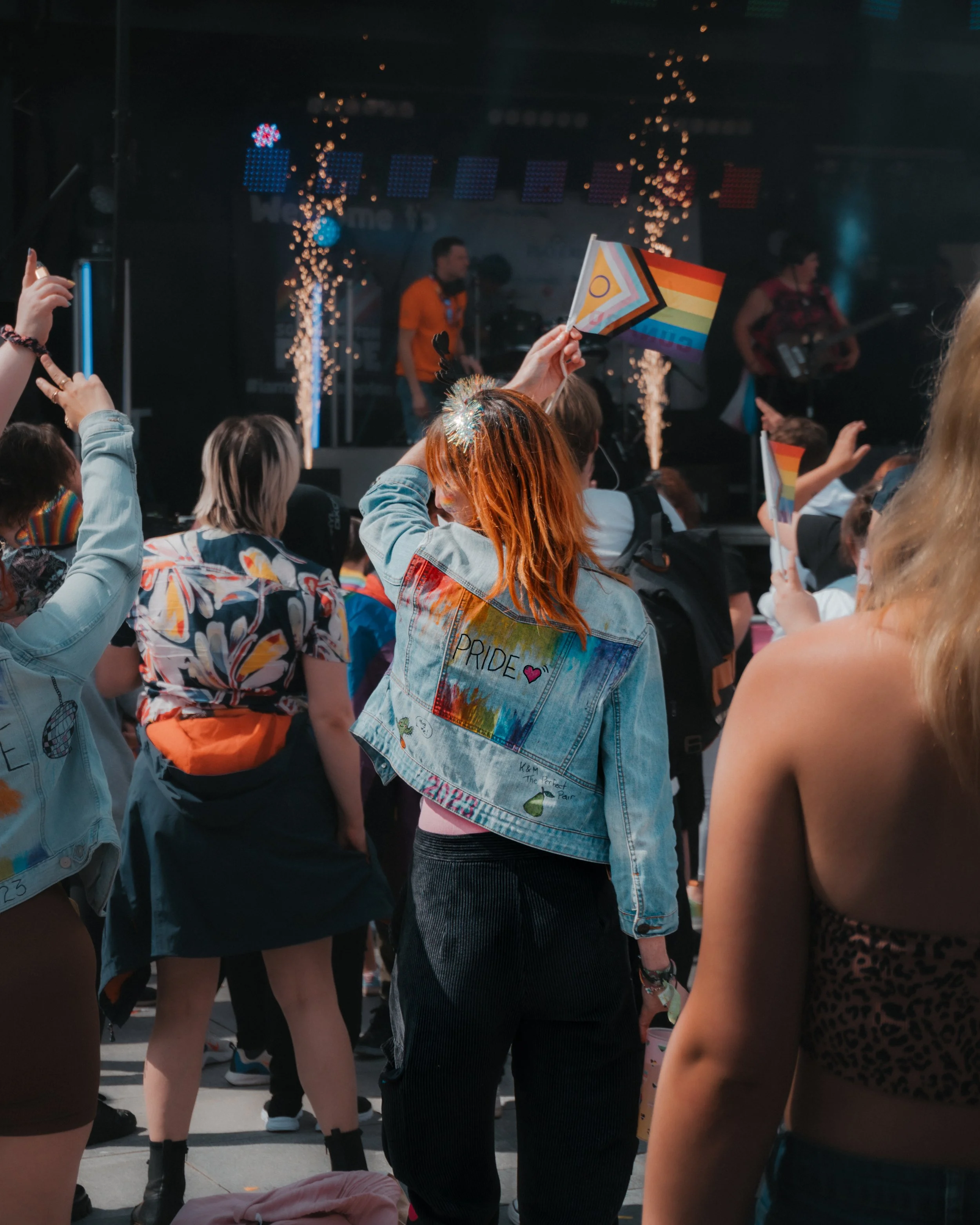 People at a Pride event, some holding rainbow flags. A person in the foreground has "Pride" on their denim jacket. There are sparkles in the air, and a stage with performers in the background.