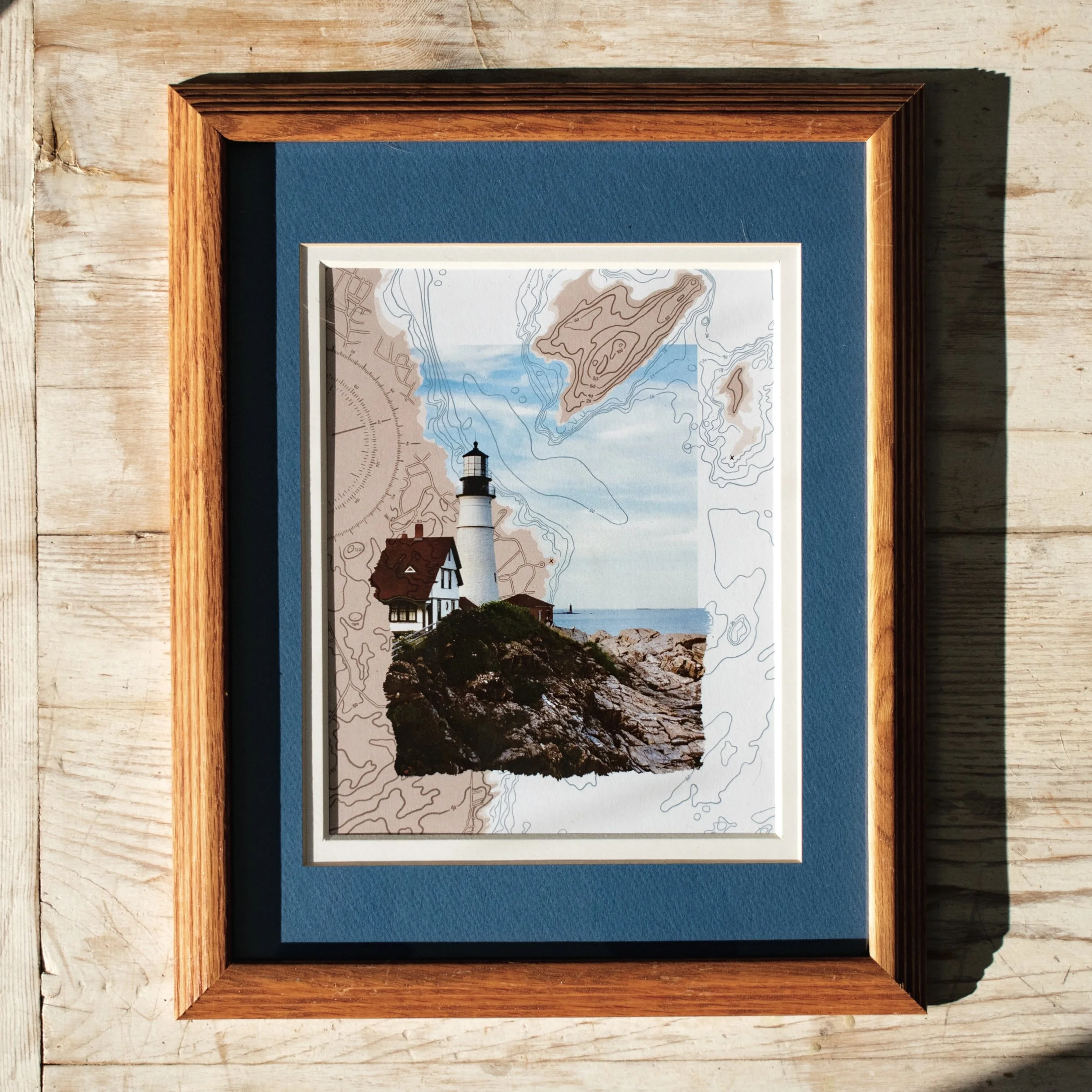 Framed artwork of a lighthouse on a rocky coast with a house, background of ocean and sky, overlaid with a topographic map.