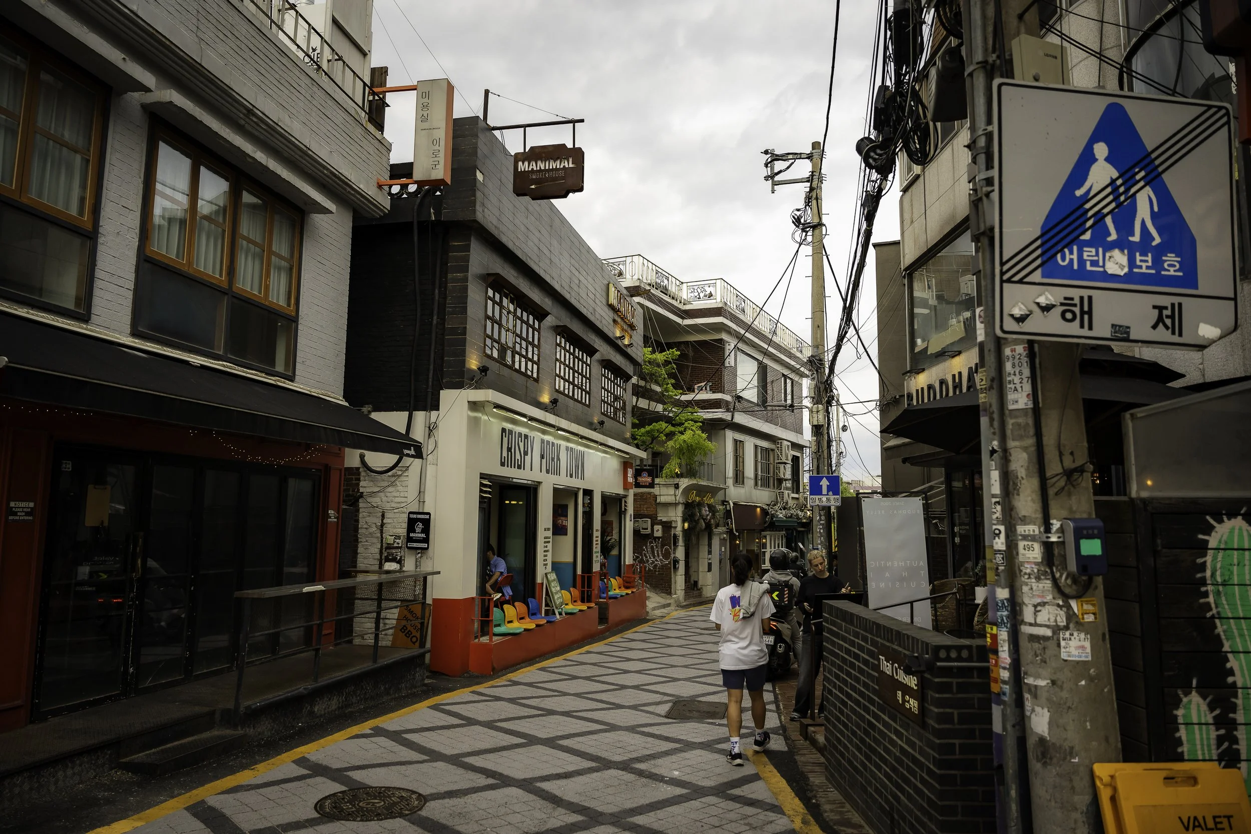 A street scene in an urban area with buildings, shops, and pedestrians. There are signs in Korean, electrical wires overhead, and a pedestrian crossing sign on a pole.