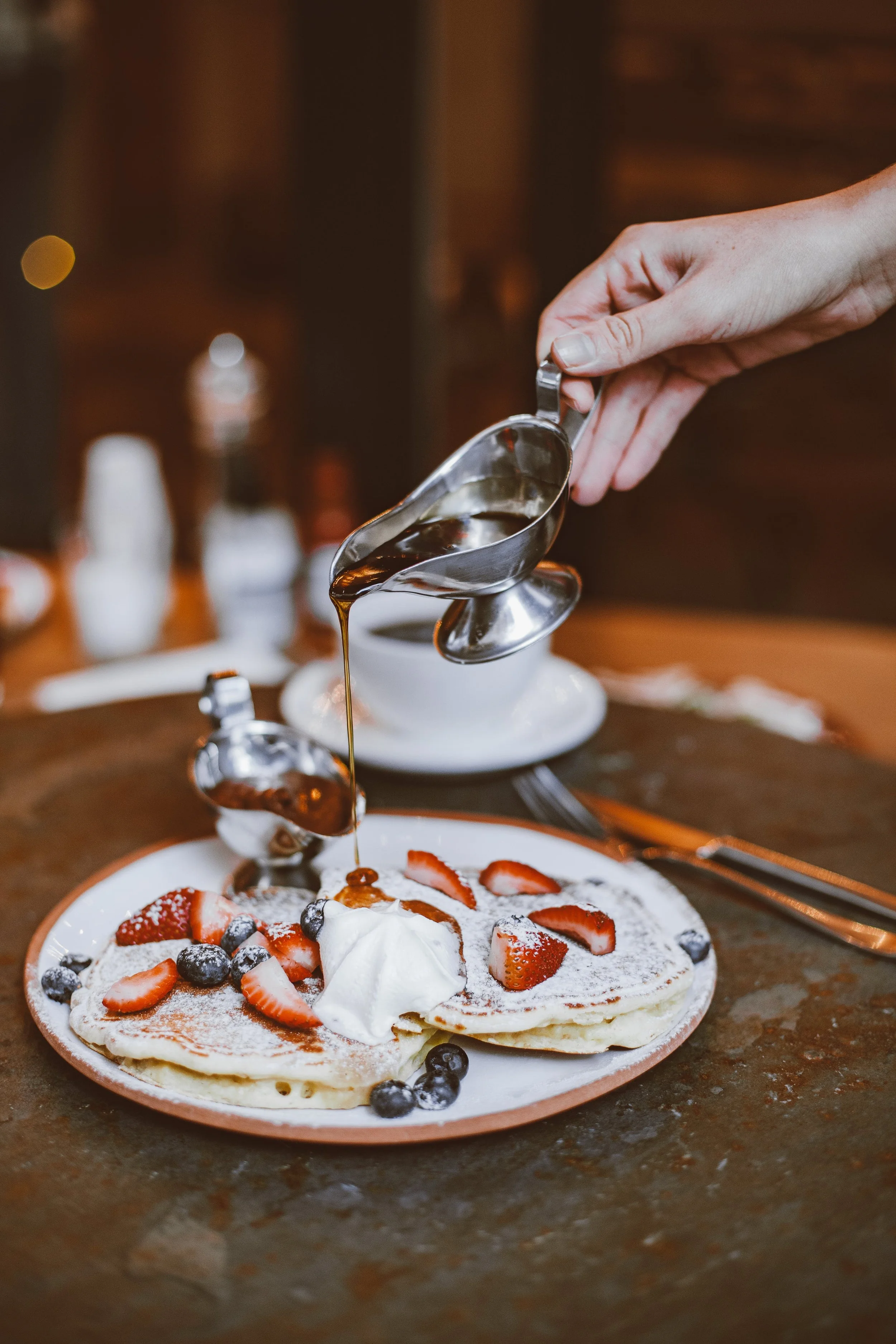 A person pouring syrup onto a plate of pancakes topped with whipped cream and fresh strawberries and blueberries.