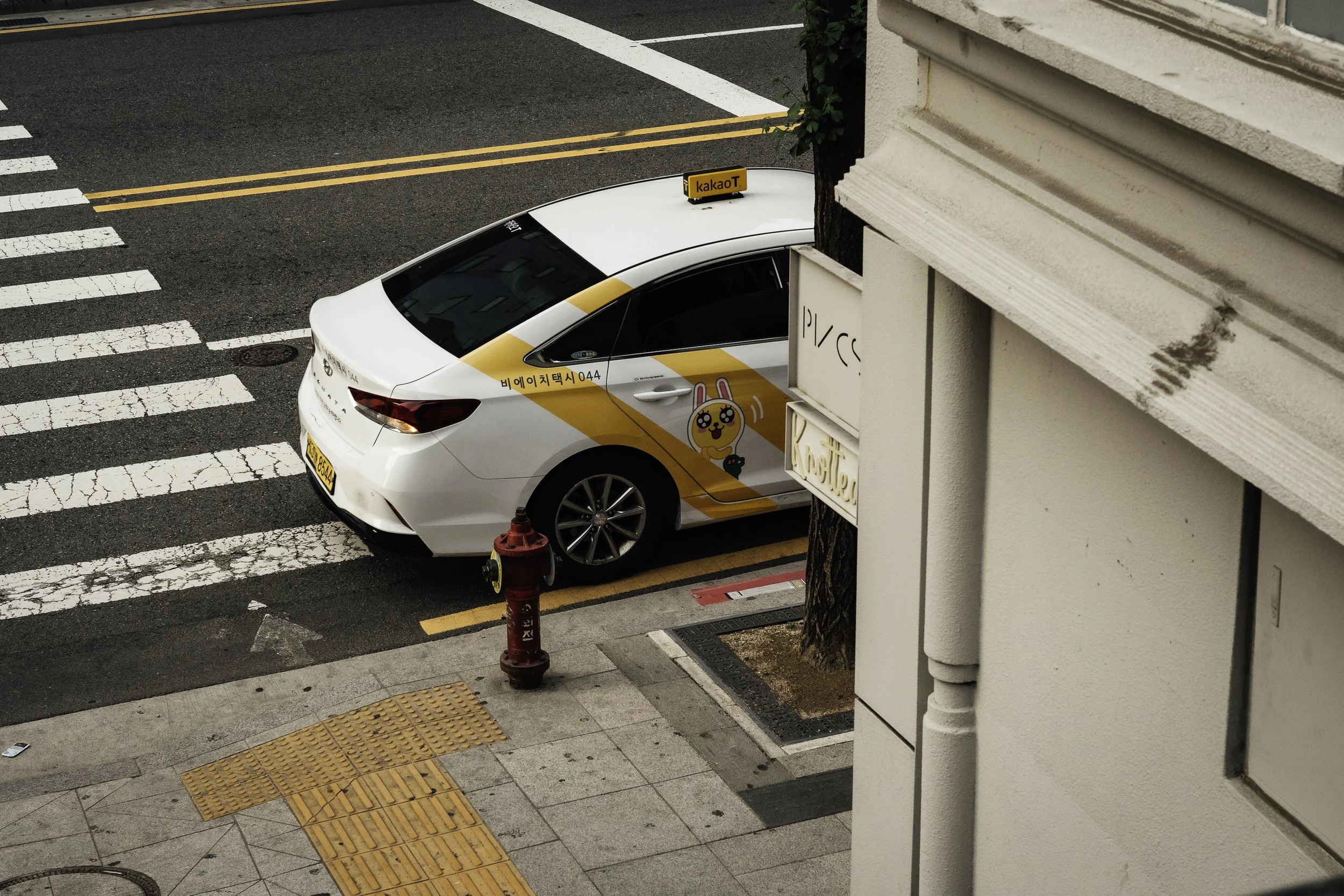 White taxi with yellow and pink cartoon character decal parked on city street near crosswalk, sidewalk, and building with signage.