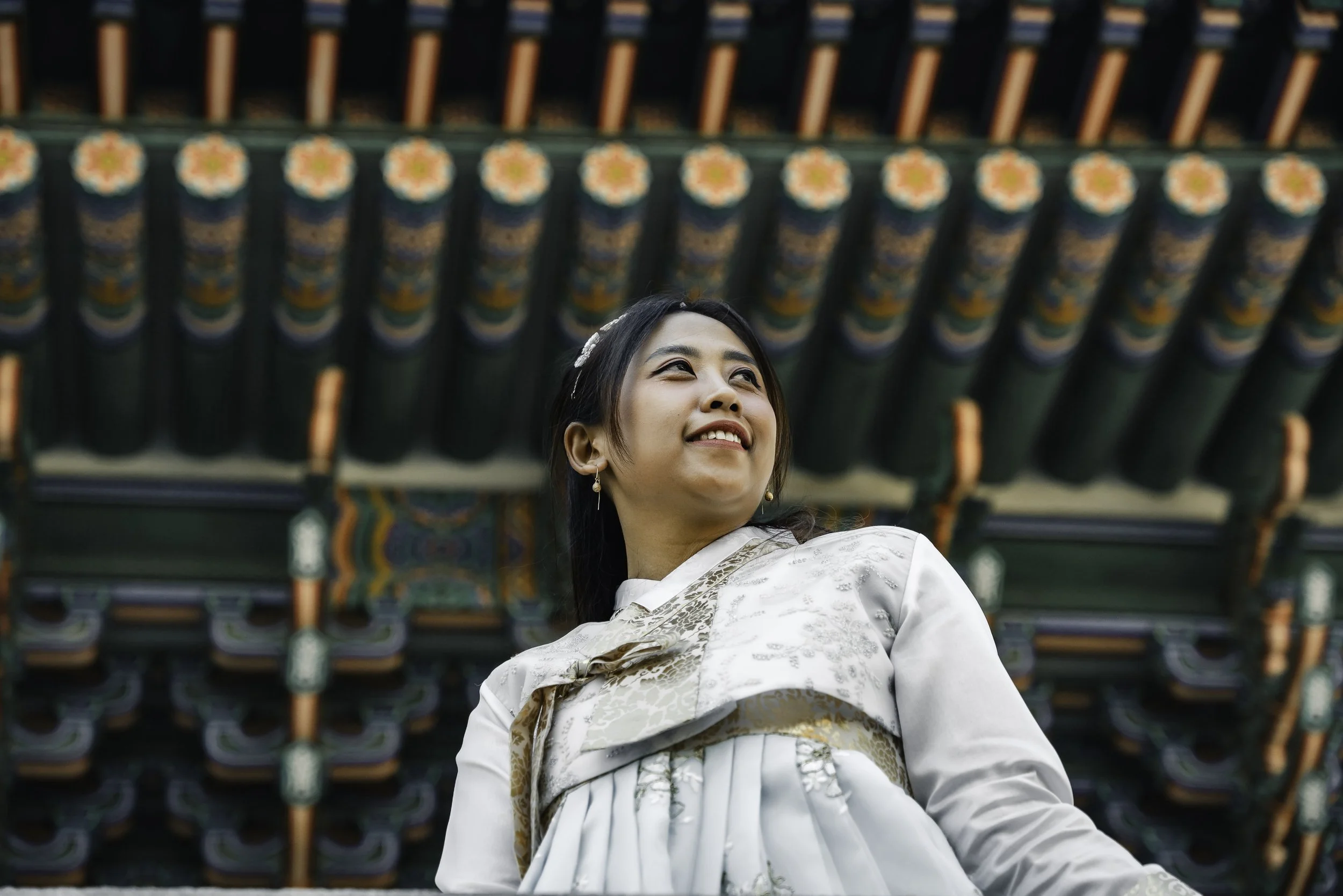 A woman in traditional Korean hanbok attire smiling and looking upward, standing in front of a colorful, ornate pavilion roof.