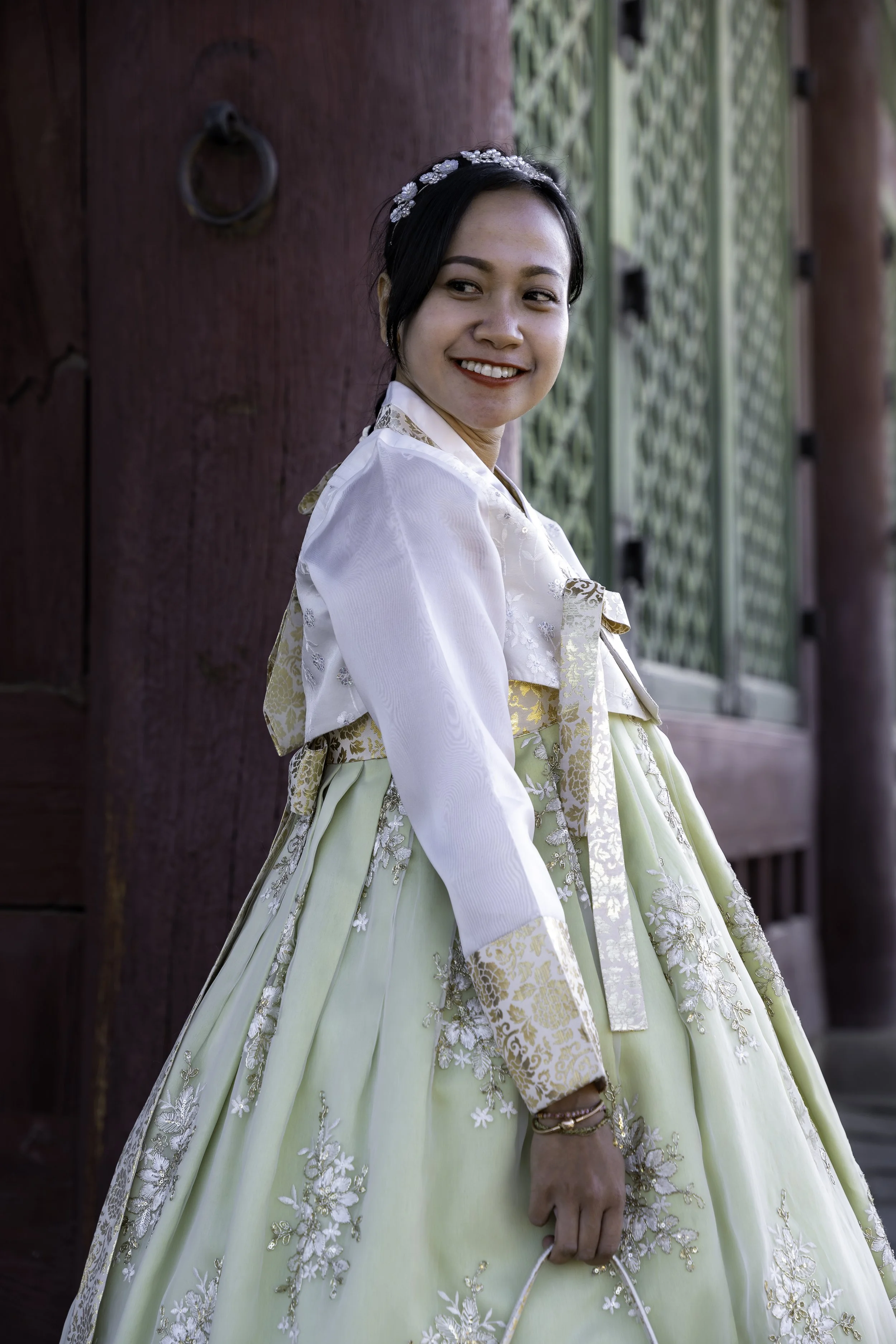 A woman in traditional Korean attire, Hanbok, smiling and holding a small handbag, standing outdoors near a wooden and green lattice structure.